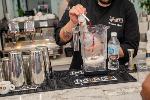 Bartender mixing a red drink in a blender at a bar counter with stainless steel shakers and a water bottle nearby.