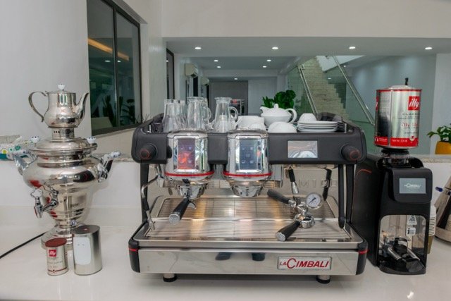 A professional espresso machine with a silver coffee grinder attached to it, a silver samovar, and various cups, glasses, and plates on a counter in a modern, well-lit interior.