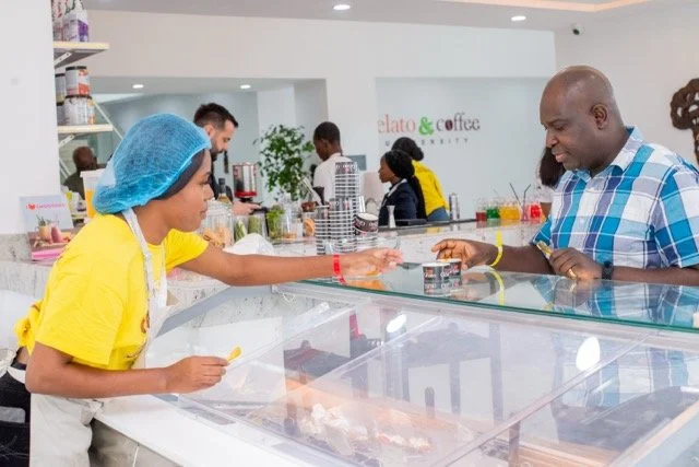 Girl in yellow shirt and blue hairnet serving ice cream to a man at a counter in a gelato shop.