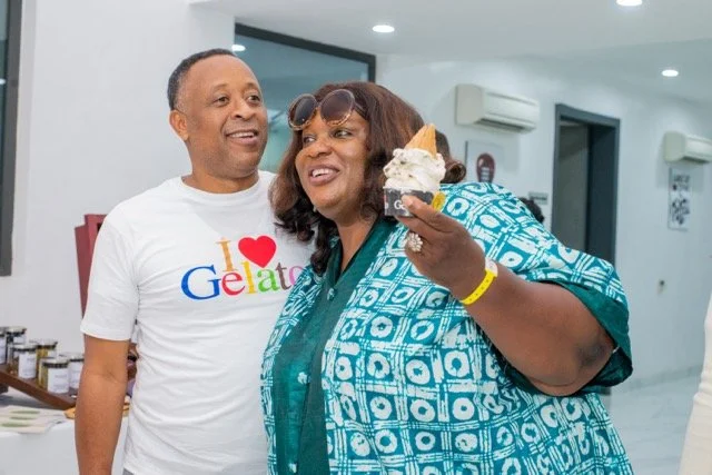 A woman enjoying ice cream inside an indoor setting, standing next to a man wearing a white T-shirt that says 'I Love Gelato' and smiling.