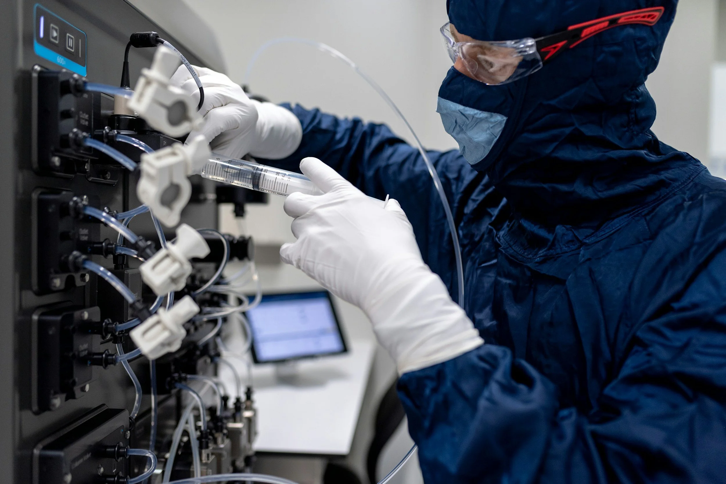 A scientist or technician working with laboratory equipment, connecting tubes and syringes on a control panel in a lab setting, wearing protective gloves and goggles.