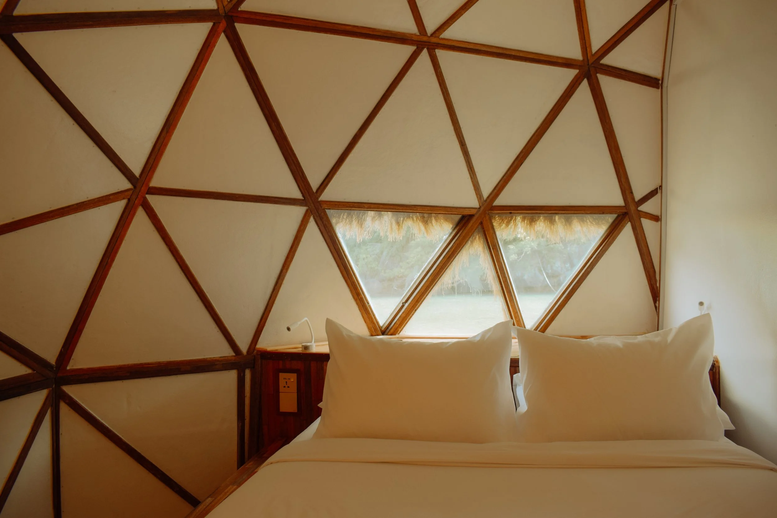 Interior of a bedroom with a geometric wooden ceiling and a large triangular window with a view of nature. A bed with white bedding is visible in the foreground.