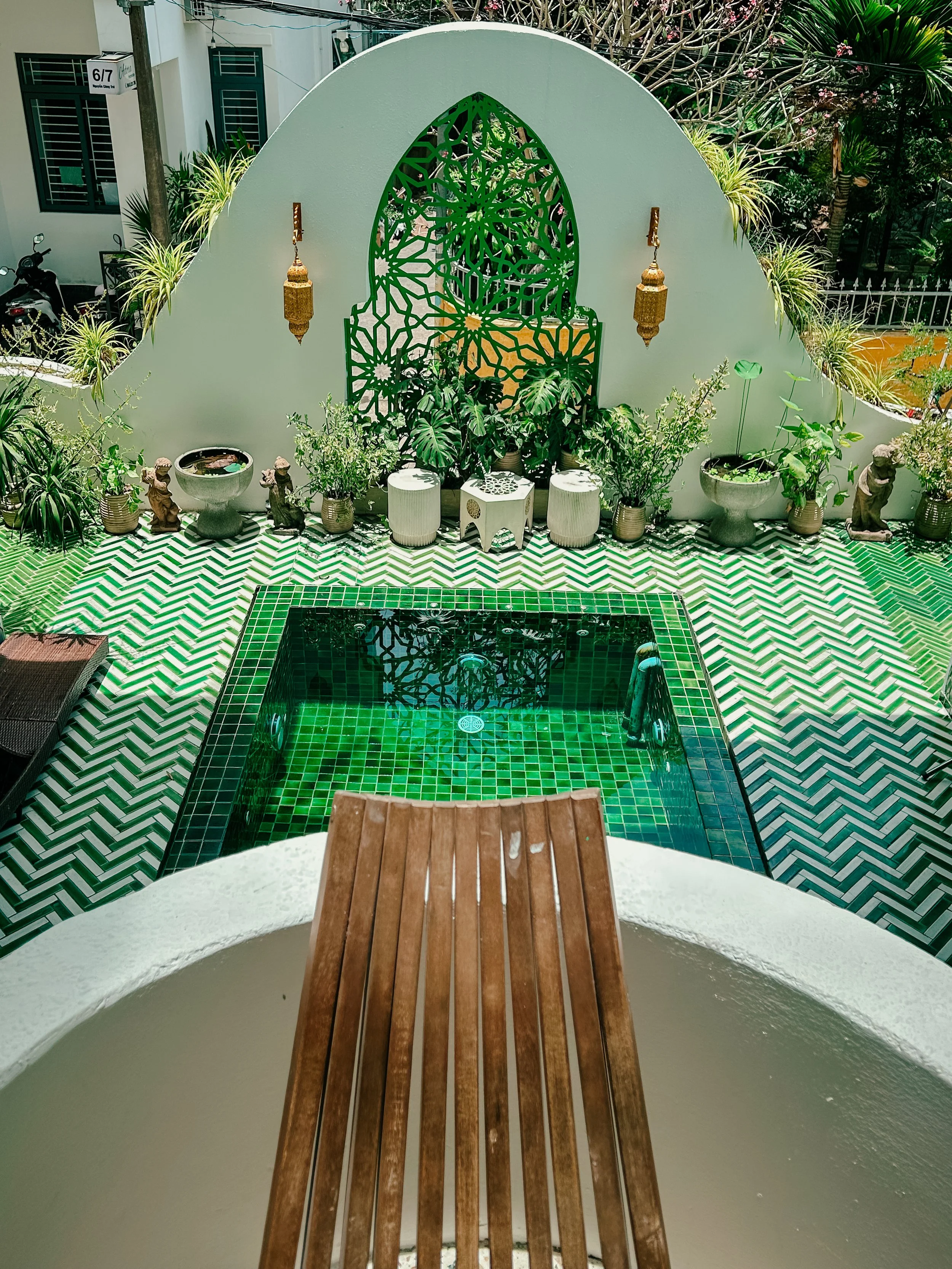 View from a balcony looking down at a small turquoise-tiled pool with a white border, surrounded by potted plants, statues, and garden decorations. A white wall with a green patterned decorative panel and hanging lanterns adorns the background.