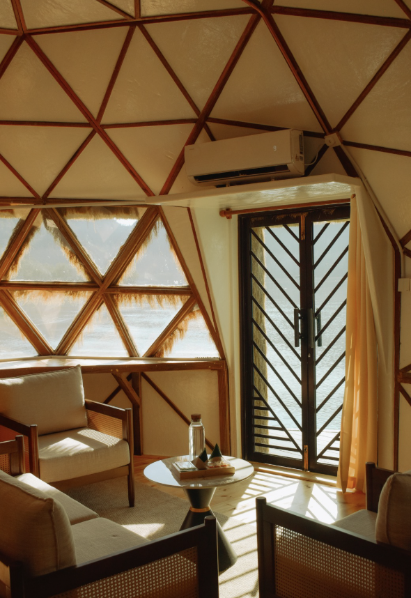 Interior of a geodesic dome with wooden framework, featuring a small seating area with beige couches and a round table near a door with a black metal grille, sunlight streaming in through triangular windows, and an air conditioning unit mounted on the ceiling.