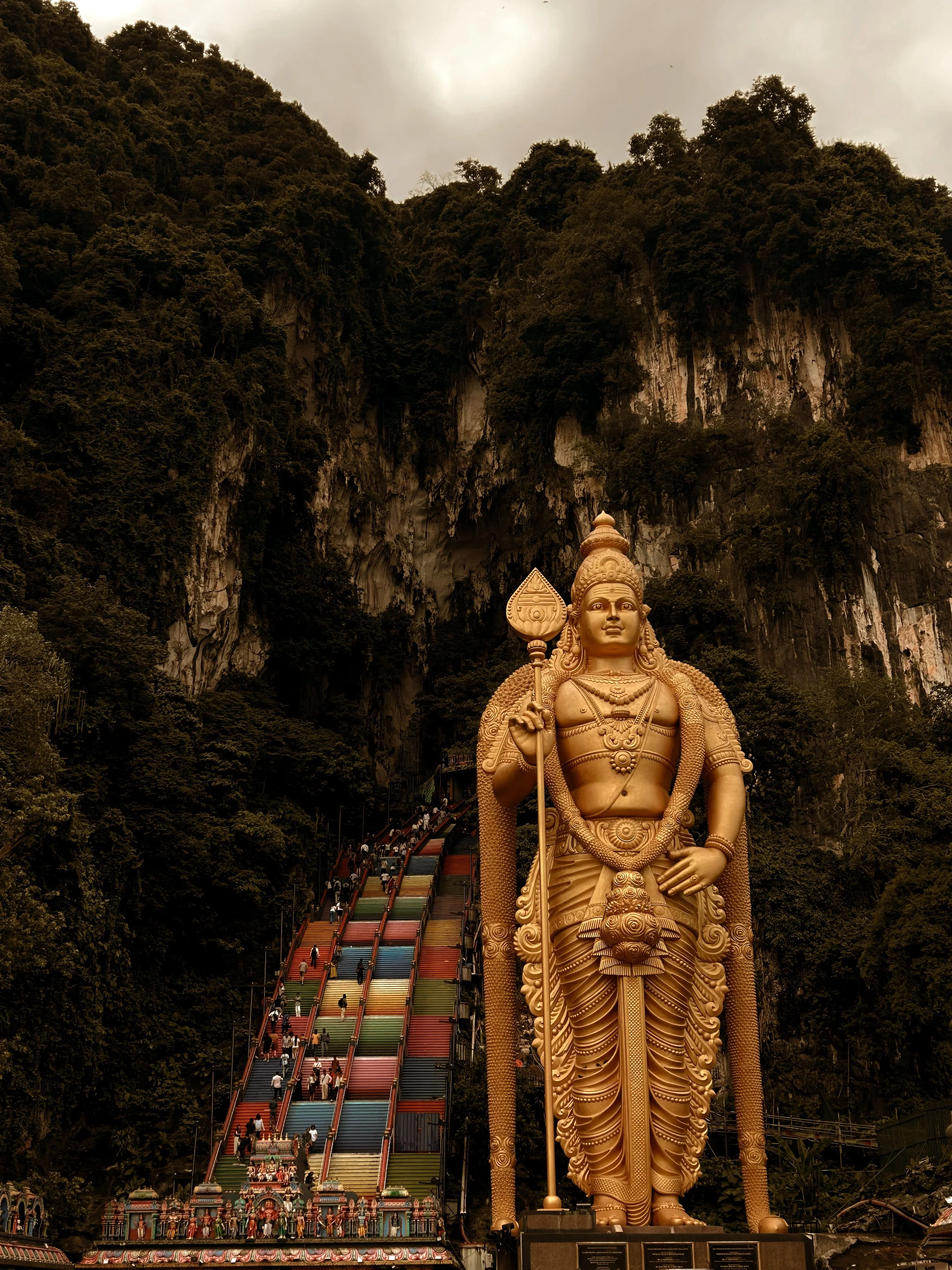 Large golden statue of Lord Murugan stands in front of a colorful staircase leading up a hill, with lush green mountain terrain and cloudy sky in the background.