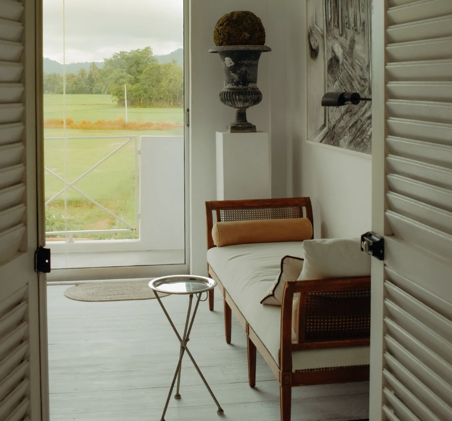 A cozy indoor space with a wooden settee, a small round table, a tall decorative urn with moss, and a view of a grassy landscape with trees and mountains through a glass door.