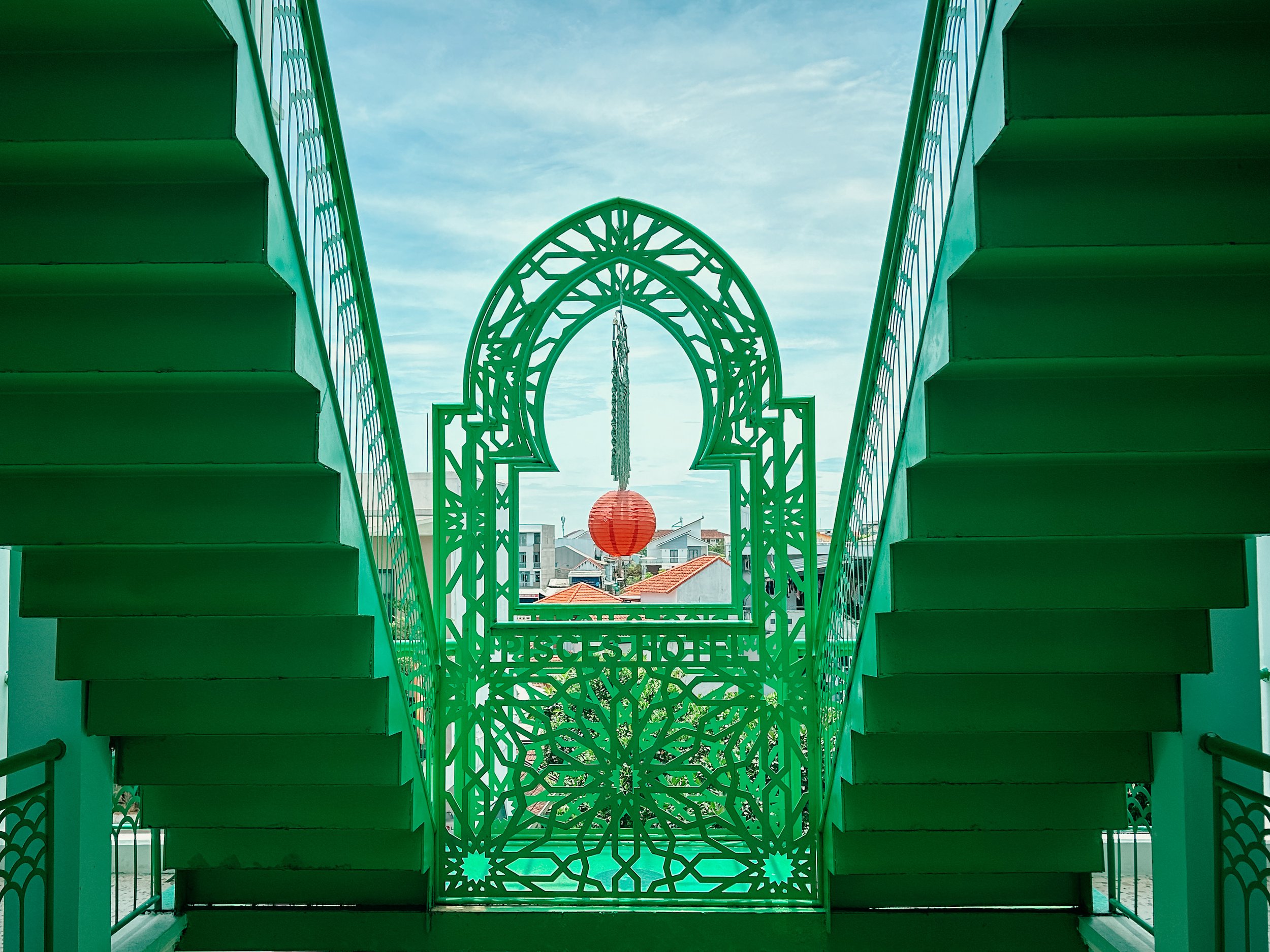 Green decorative iron gate with geometric and floral patterns, flanked by green stairs on both sides, with a view of a cityscape and a cloudy sky in the background.