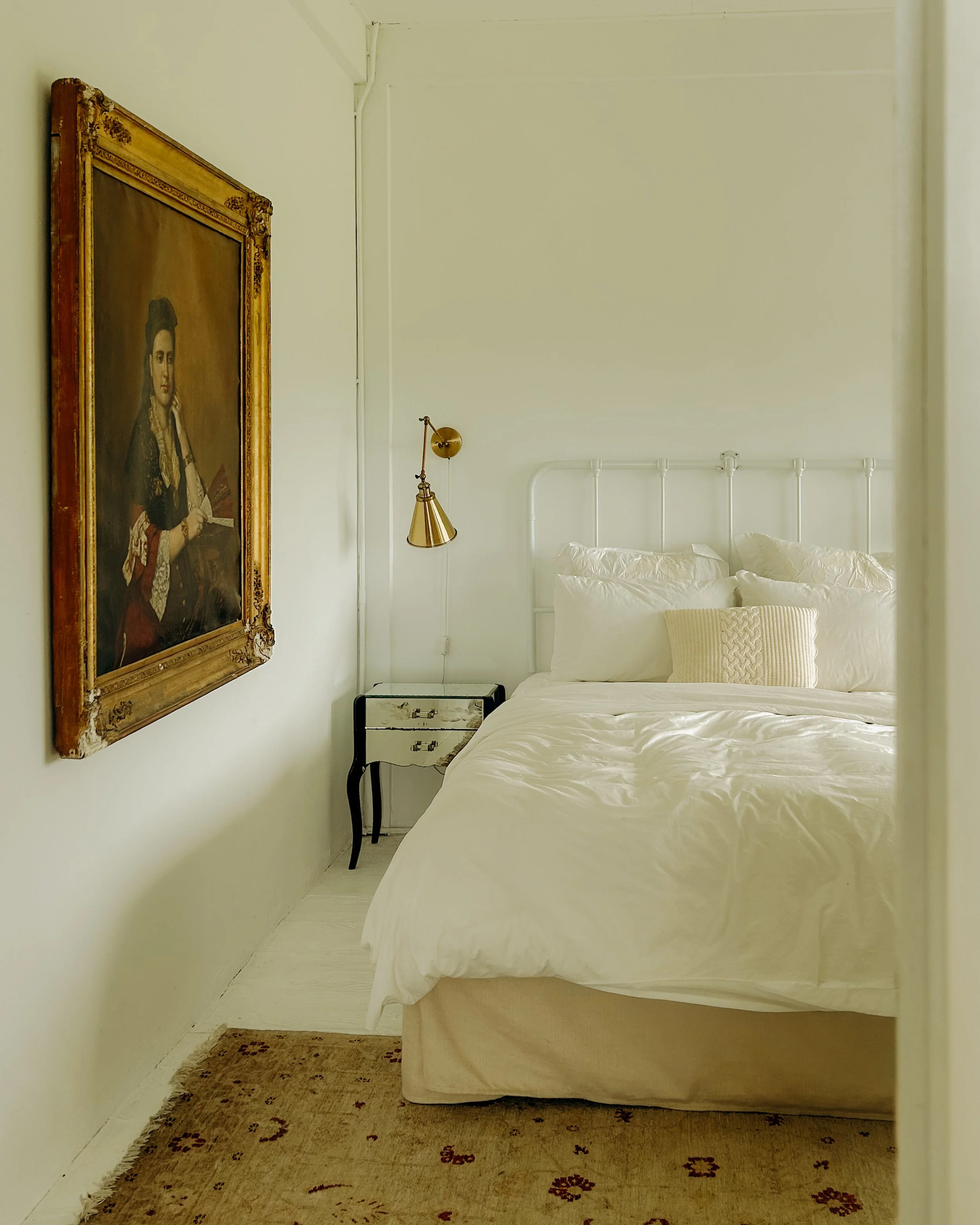 A simple bedroom with a white bed, pillows, a framed portrait on the wall, a gold wall-mounted lamp, and a beige carpet on the floor.