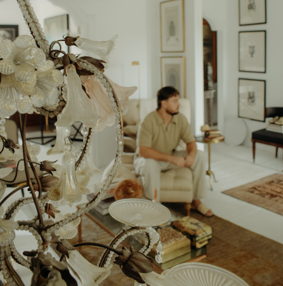 Living room with a man sitting on a cream-colored armchair, surrounded by framed pictures and a side table with books. In the foreground, a detailed chandelier with glass and bead decorations is visible.