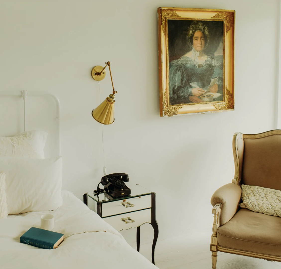 A cozy bedroom corner featuring a white bed with pillows, a glass nightstand with a vintage rotary phone, a black and gold lamp, a framed oil portrait of a woman on the wall, and a beige upholstered armchair.