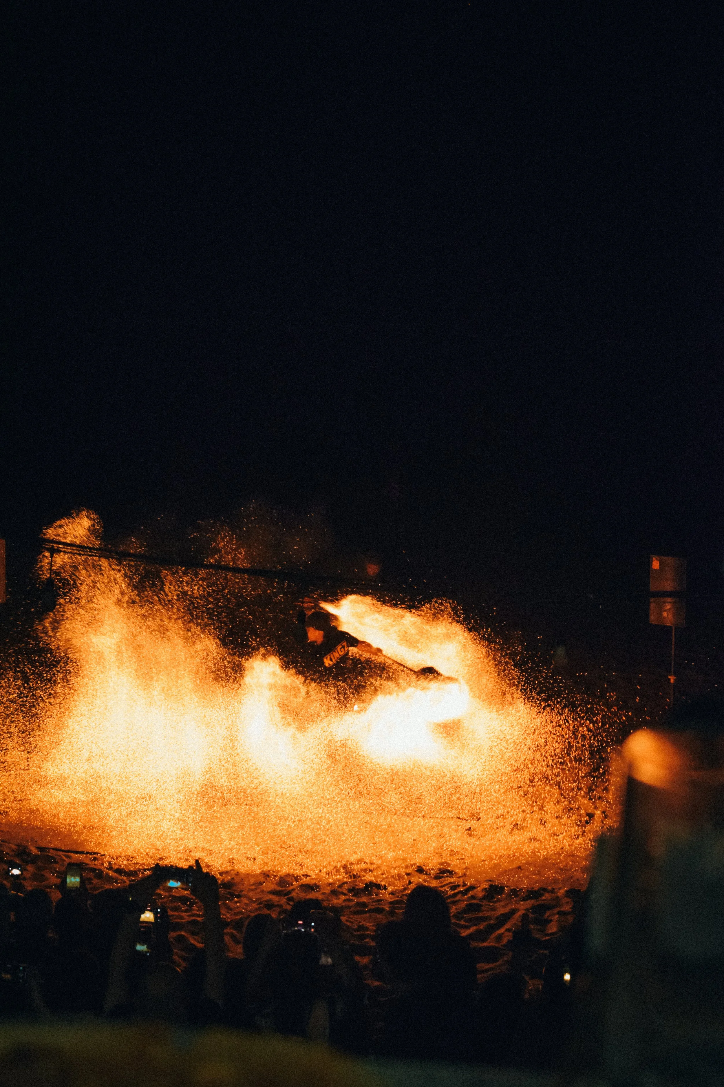 People gathered at night watching a large fire or blaze with sparks flying into the air, some taking photos with their phones.