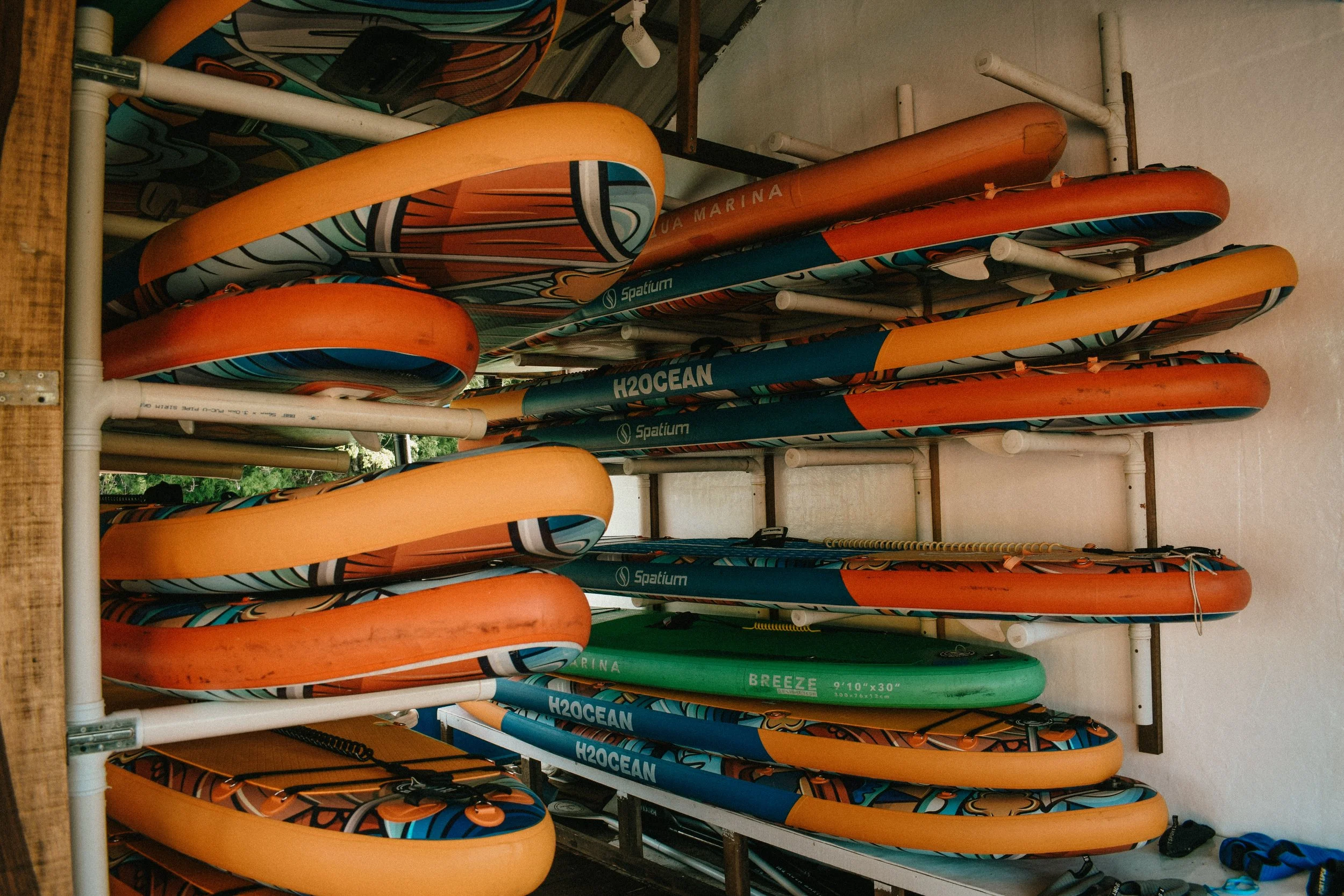 Multiple orange, blue, and green paddles stacked on a shelf, with some labeled with brand names and sizes.