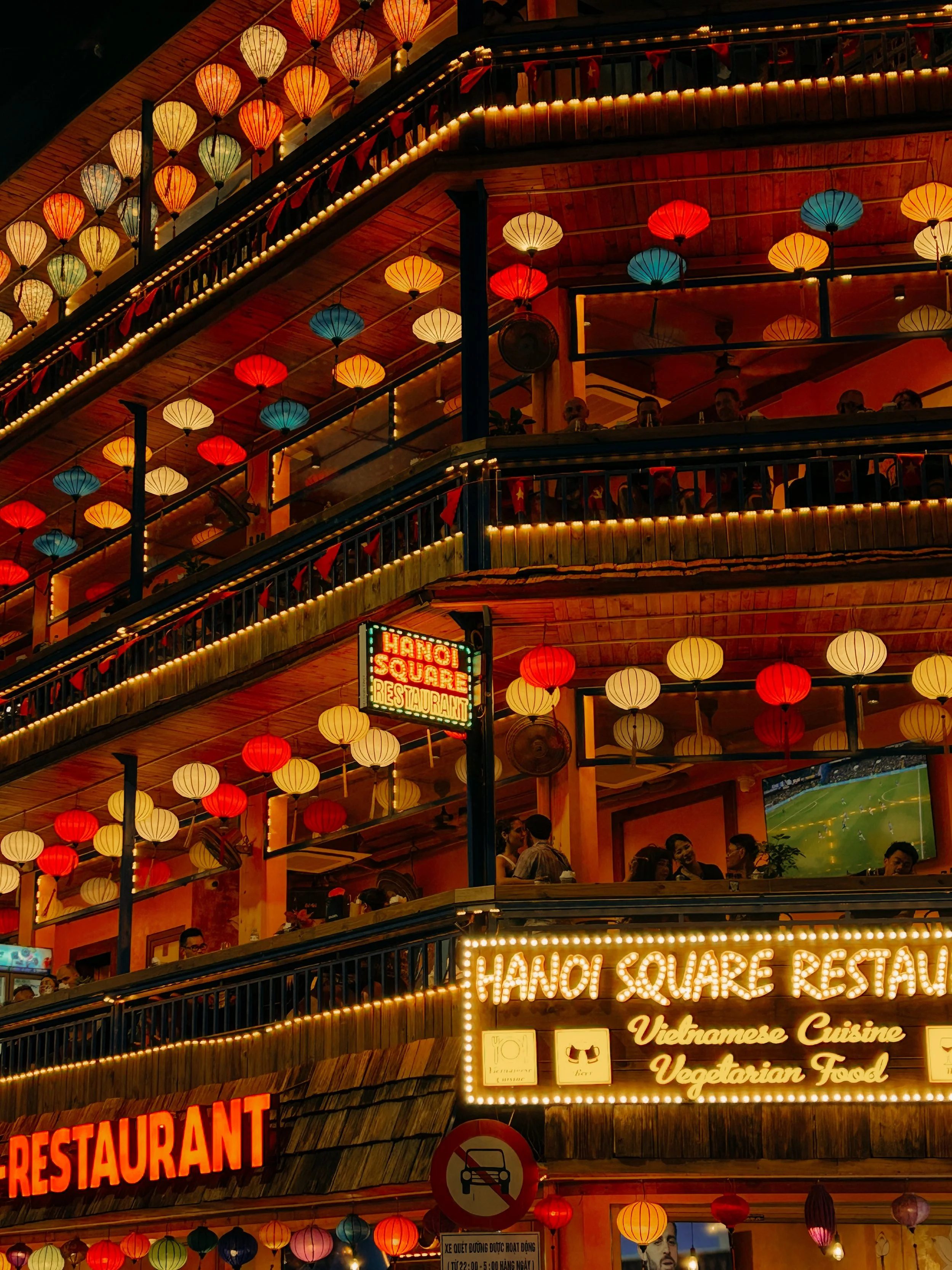 Multiple floors with colorful hanging lanterns, illuminated signs for 'Hanoi Square Restaurant' and Vietnamese cuisine, glowing lights, and people dining at night.