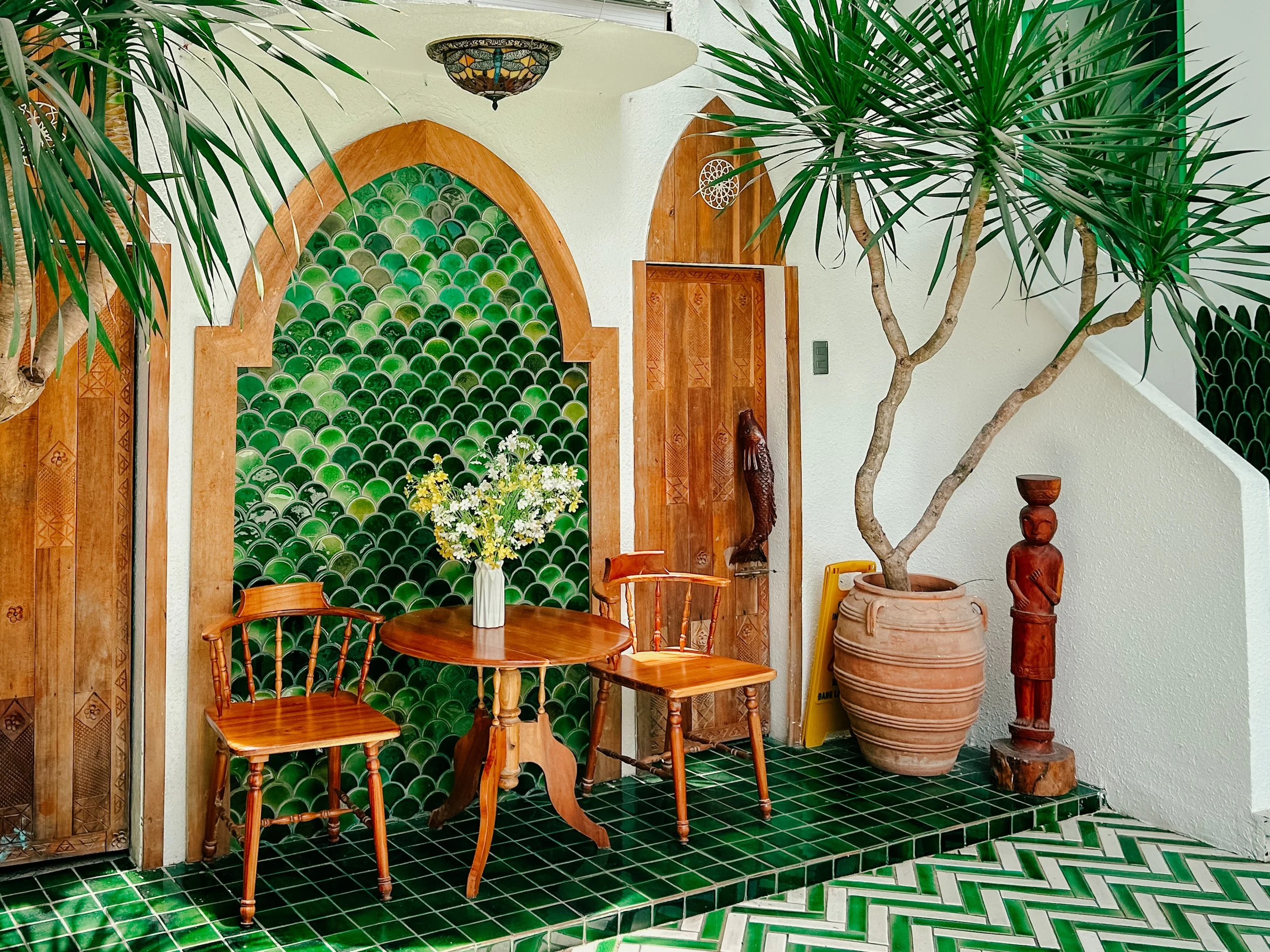 Interior of a room featuring green tiled floor and wall, wooden furniture including a round table with a white vase of white flowers, chairs, tall potted plants, and decorative wooden statues.