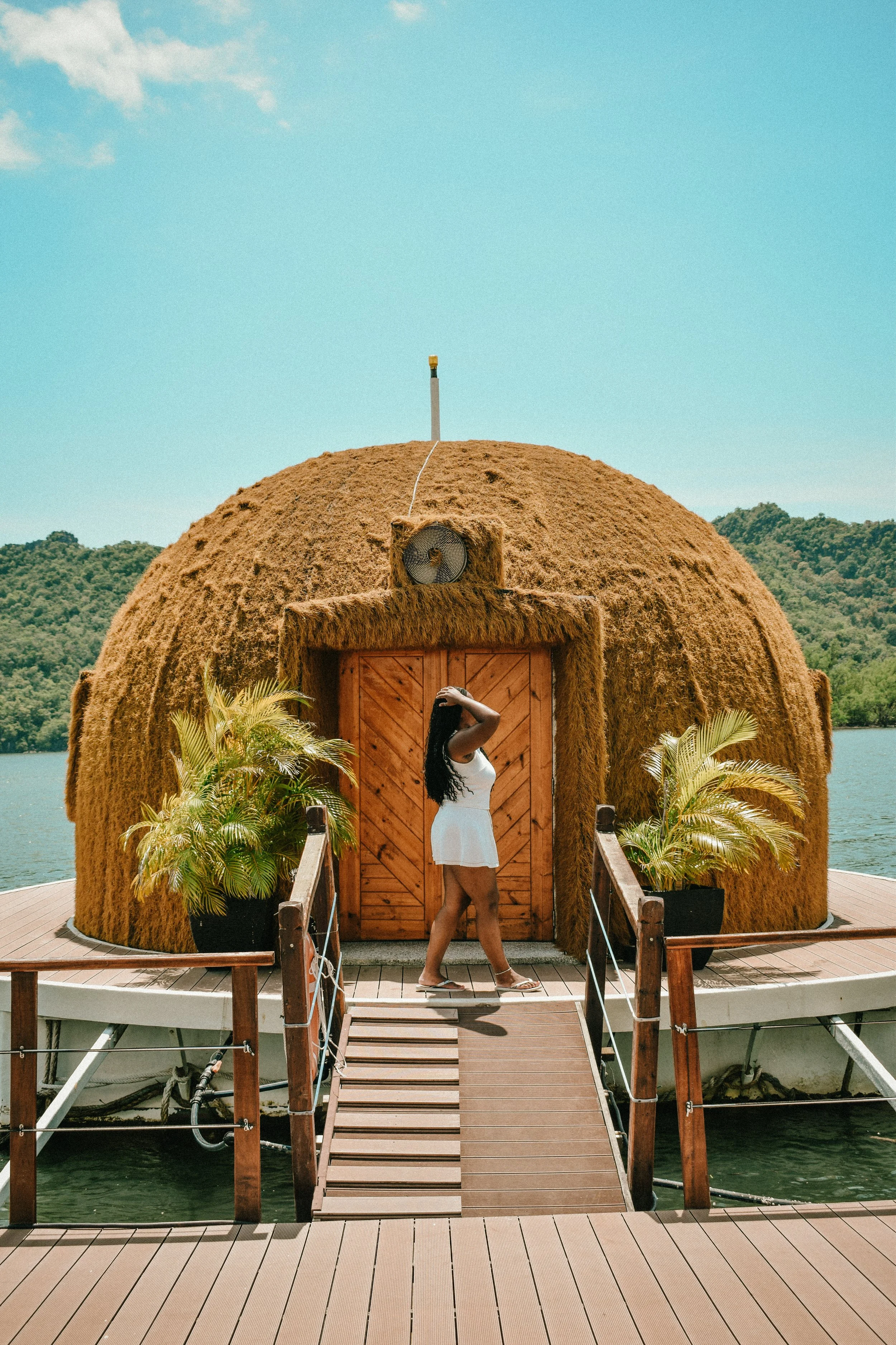A woman in a white dress, standing on a boat dock, taking a photo of a hut made of hay with a wooden door, situated on a body of water with green hills in the background.