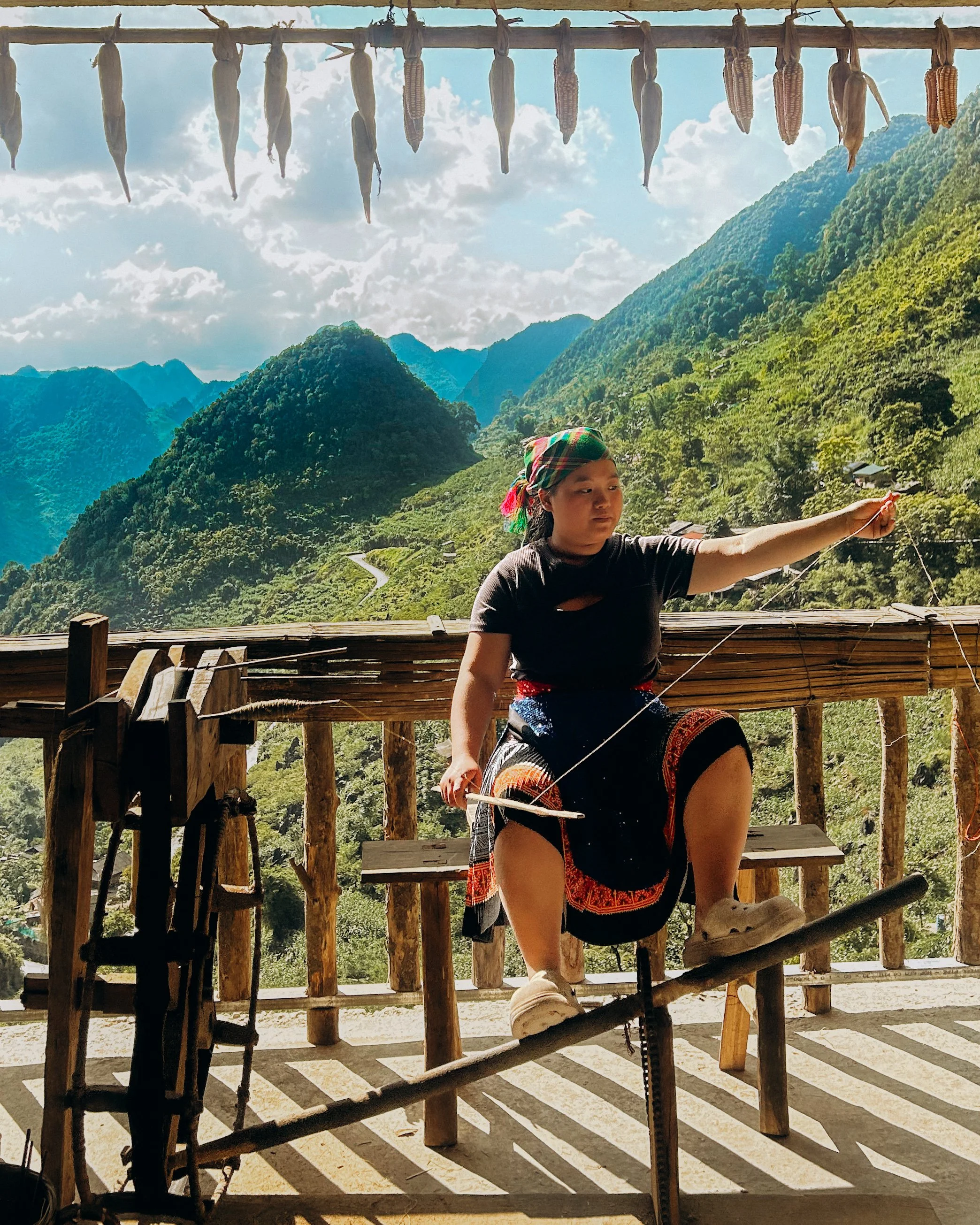 A young girl wearing traditional clothing and a colorful headscarf, balancing on a wooden log above the ground while holding a string. She is in a mountainous, rural area with lush green hills and scattered houses in the background. The scene is shaded by a wooden structure, with corn and other crops hanging to dry overhead.