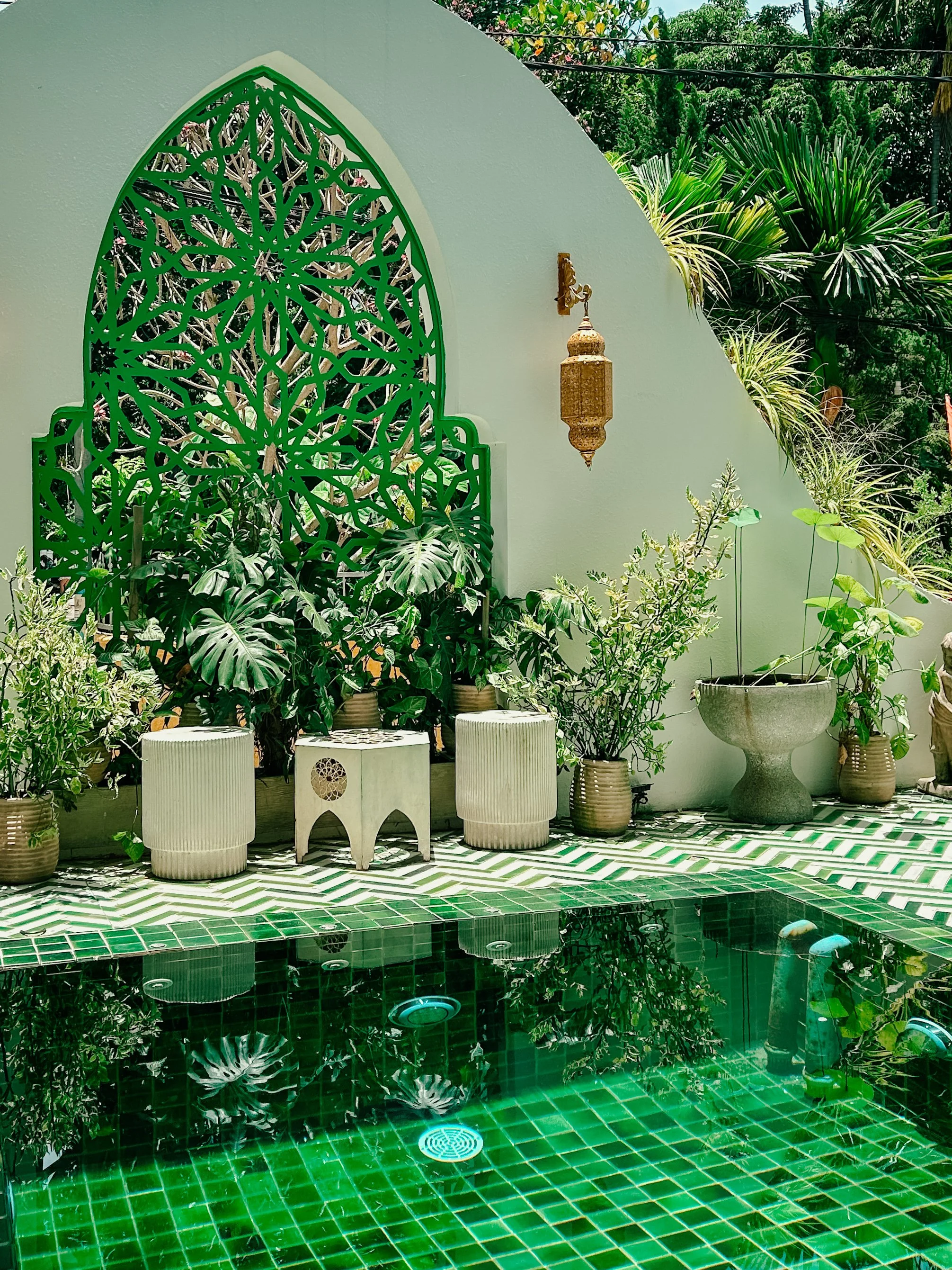 A decorative outdoor patio area with white pottery planters with green plants, a white wall with a green decorative window, and a swimming pool with green mosaic tiles reflecting plants and blue sky.