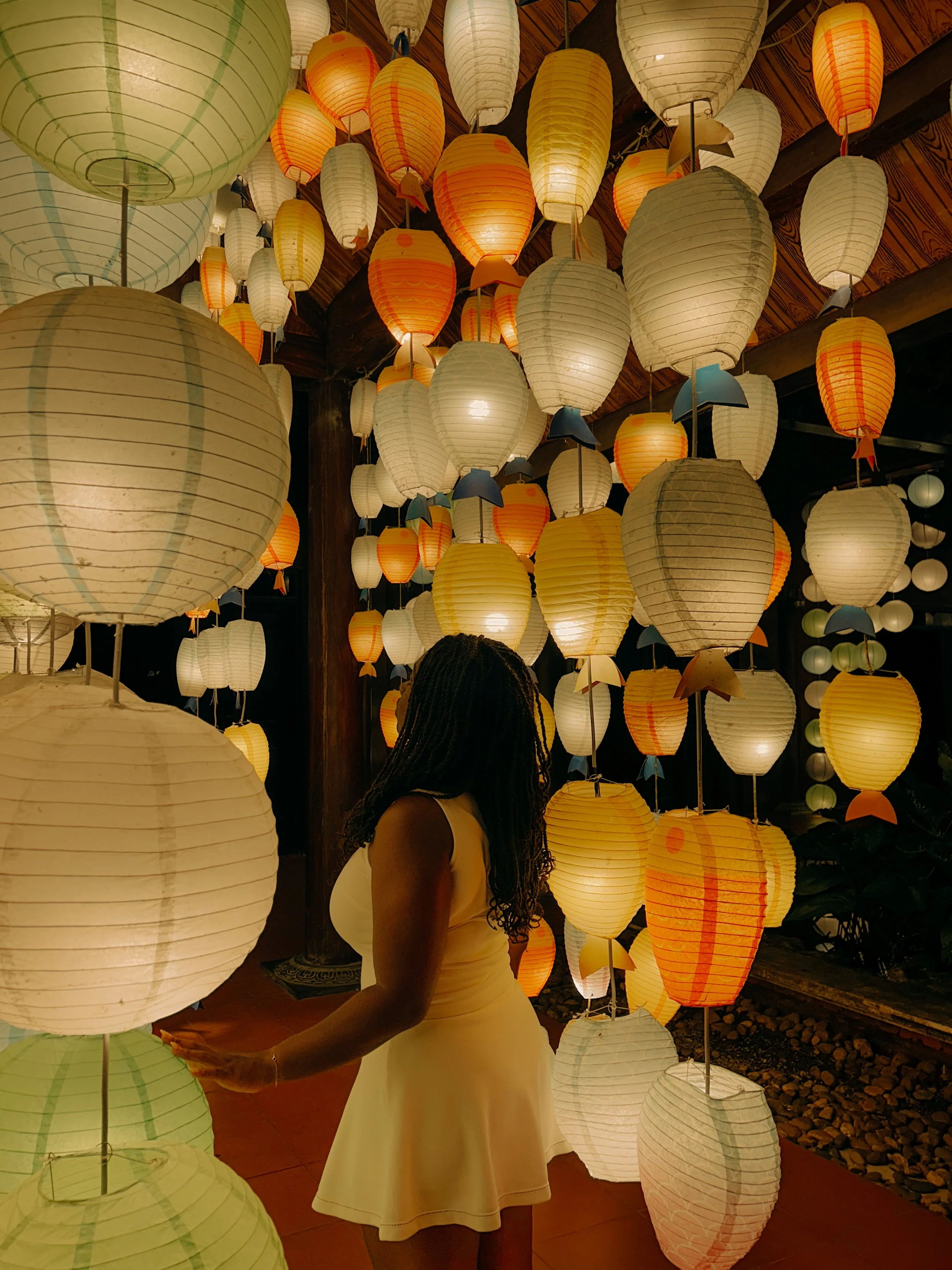 A woman in a beige dress standing among hanging paper lanterns in various shades of white, yellow, and orange, with some holding umbrellas, creating a colorful and festive scene.