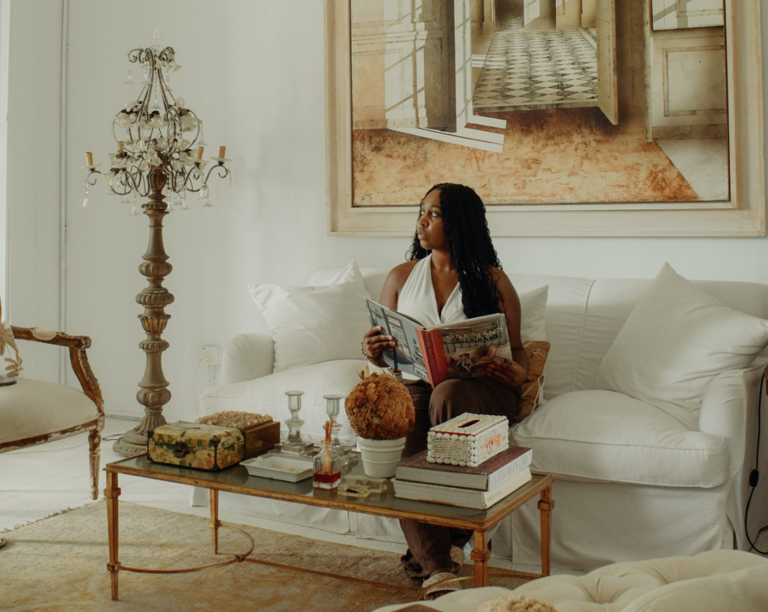A woman with long black curly hair sitting on a white sofa, holding a magazine, in a well-decorated living room with a coffee table and a large painting on the wall.