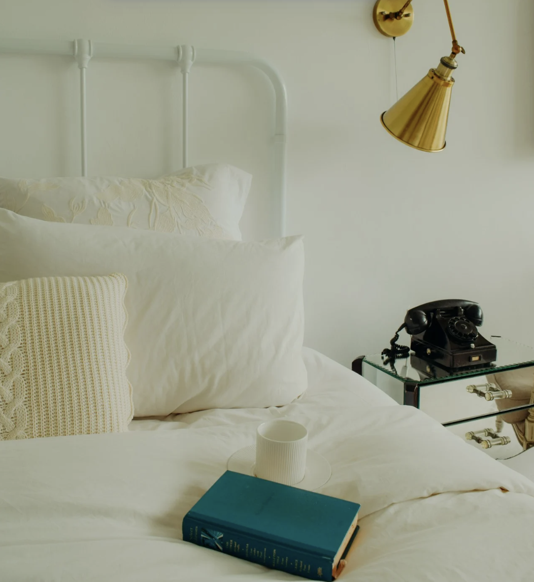 A cozy bedroom with white bedding, pillows, and a knitted cream pillow. A blue book and a white cup are on the bed. A vintage black rotary phone is on a mirrored side table. A gold wall-mounted lamp hangs above, against a cream-colored wall.