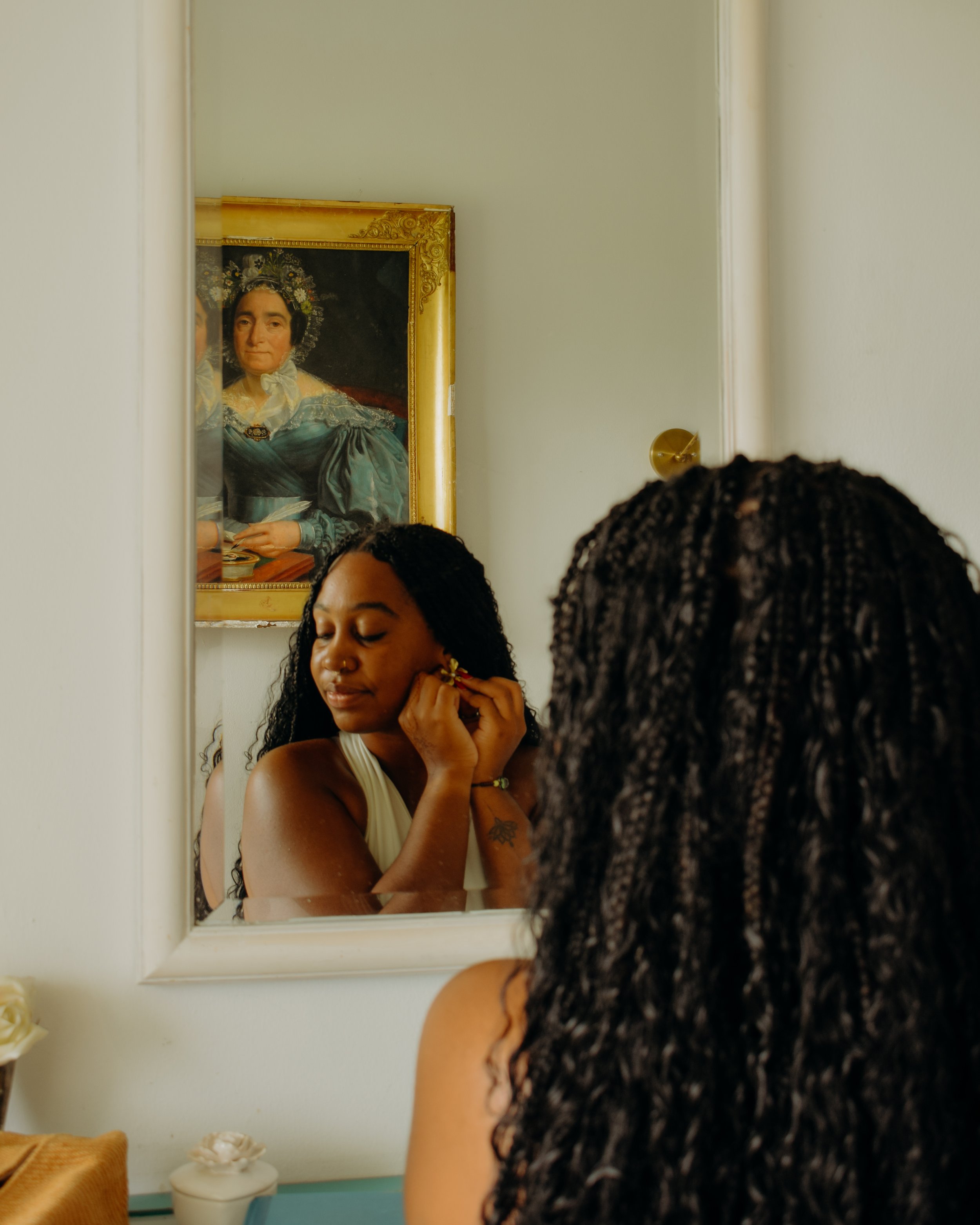 A woman with long curly black hair is putting on earrings in front of a mirror. The reflection shows her face and part of her arm. Behind her, there is a framed portrait of a woman dressed in historical clothing hanging on the wall.