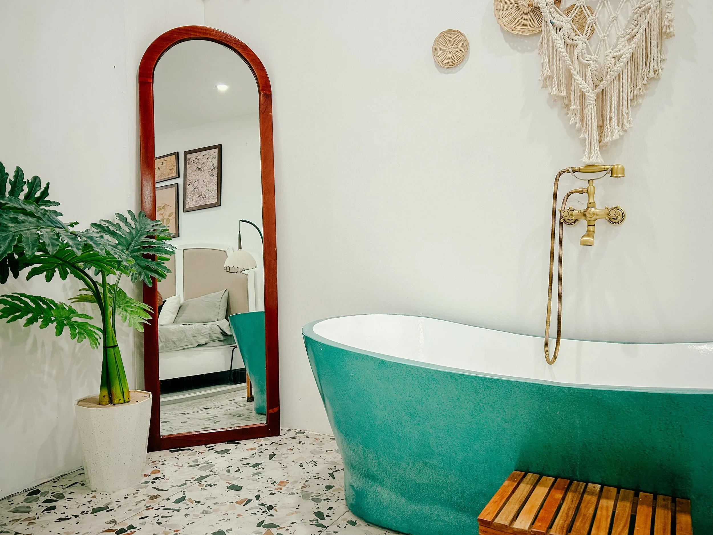 A bathroom with a turquoise bathtub, a gold shower faucet, a large mirror with a wooden frame, and decorative wall art made of woven baskets and macramé hanging on the white wall. A large green plant in a white pot is also present.