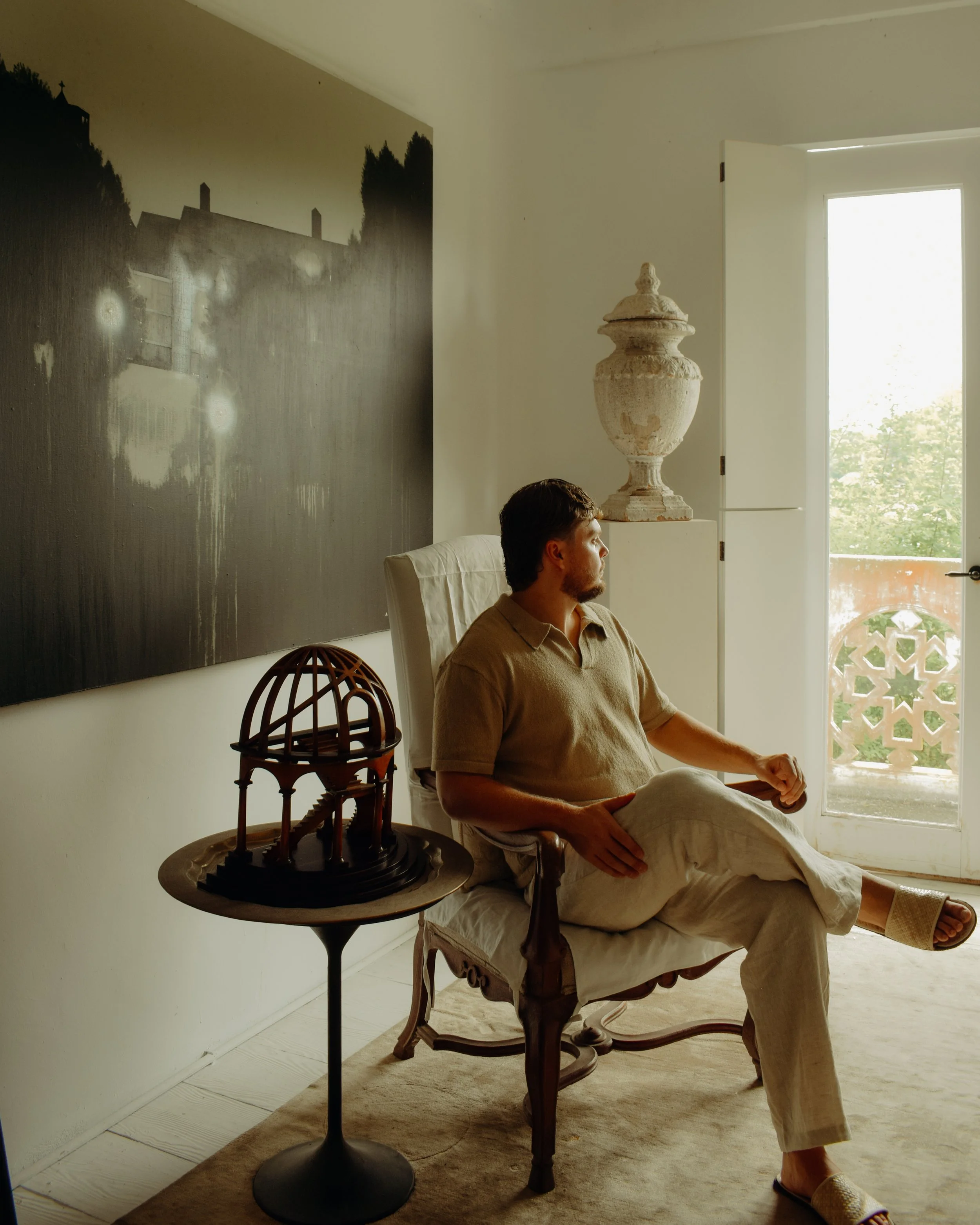 Man sitting in a vintage wooden chair, wearing a beige shirt and light-colored pants, with a relaxed posture, inside a room with a large open window. A decorative urn is on a stand beside him, and a framed black-and-white photograph of a house and trees is on the wall behind him. A small table with a wooden model of a building is in the foreground.