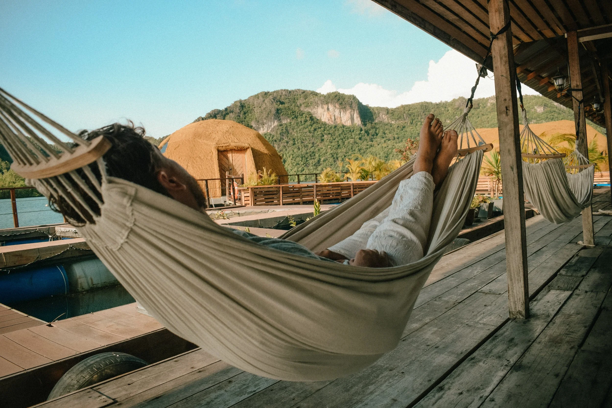 A person relaxing in a hammock on a wooden deck, with mountains and a thatched hut in the background, under a bright blue sky.