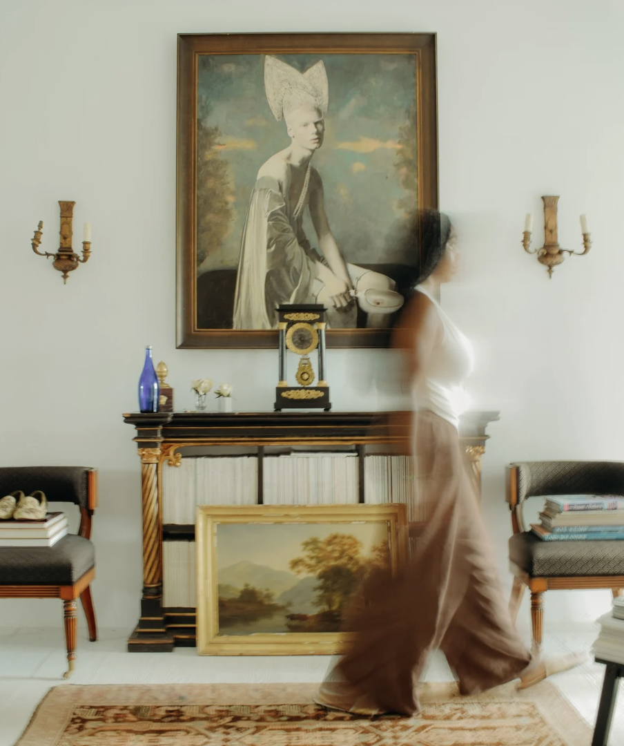A woman wearing a white sleeveless top and brown wide-leg pants walking in a living room with framed paintings, a large portrait of a woman with an elaborate headdress, two wall-mounted candle holders, and a decorative fireplace with books and objects on top.