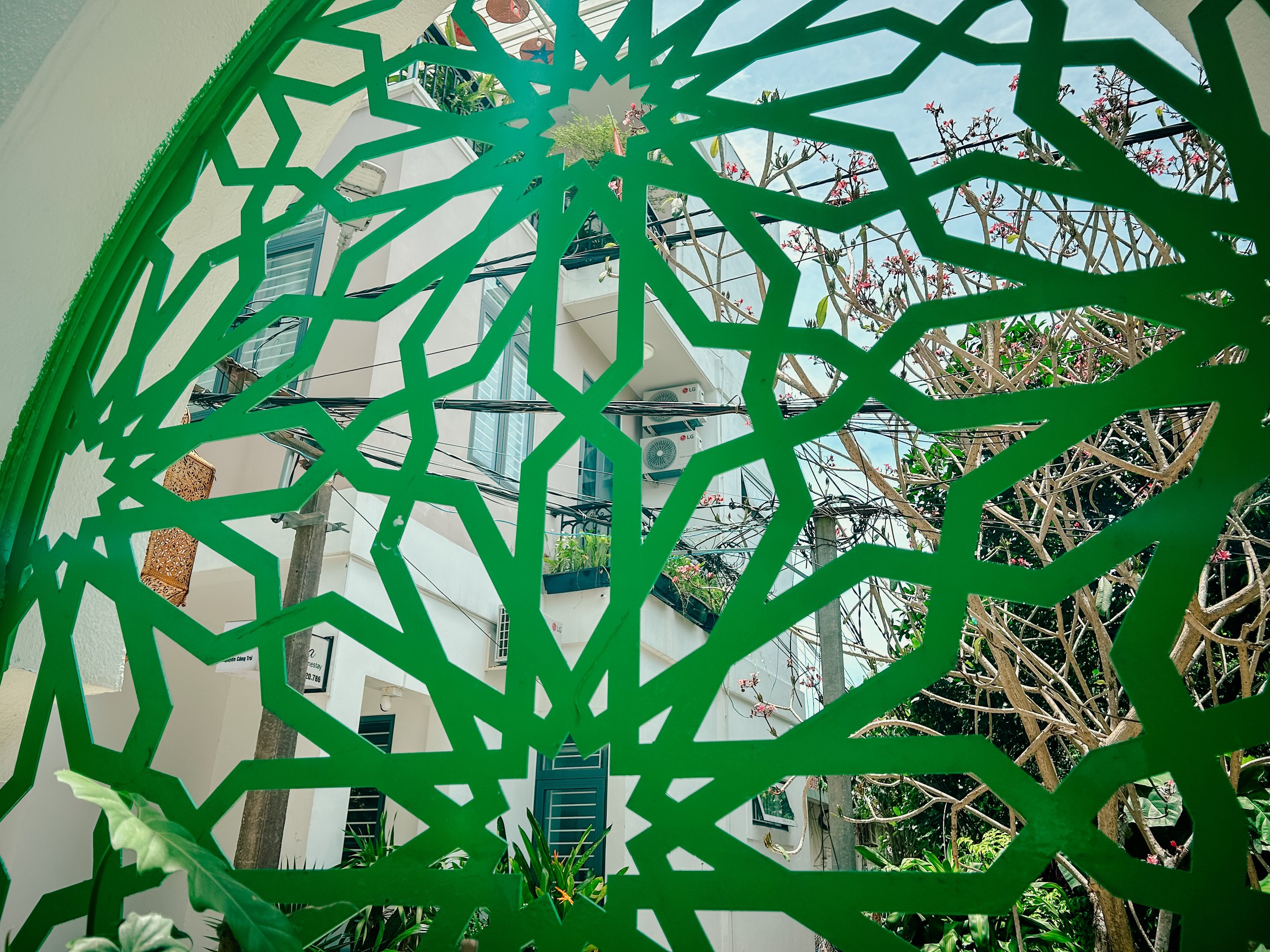 View through a decorative green metal grille with a geometric starburst pattern, revealing a residential building with air conditioning units, windows, and flowering trees in the background.