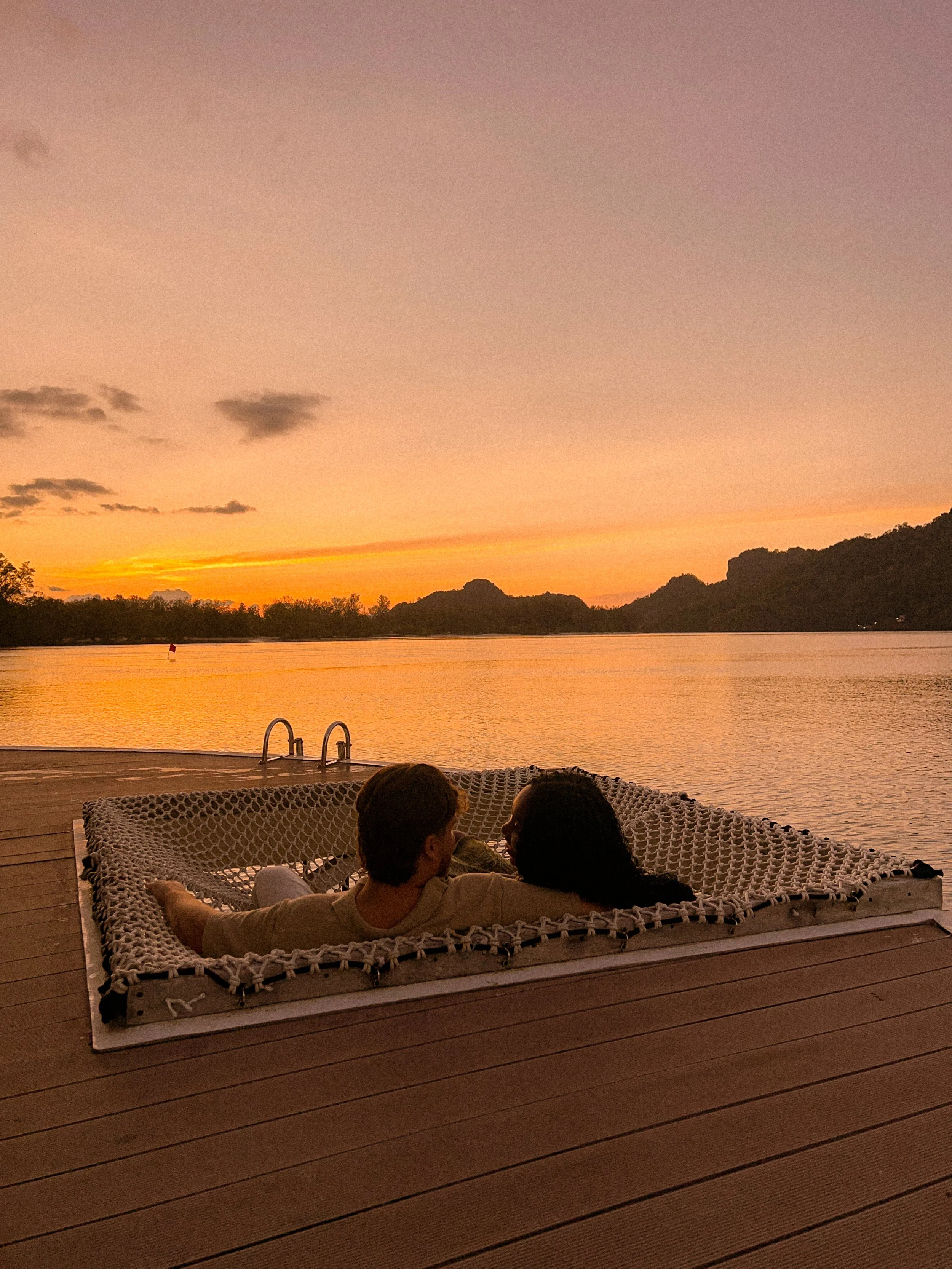 Two people are relaxing on a woven outdoor lounge chair at the edge of a lake during sunset. The sky displays shades of orange and pink, with distant hills and trees along the shoreline.