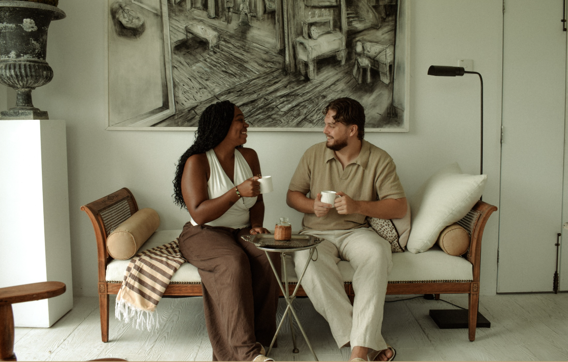 A man and woman sitting on a white sofa in a living room, sharing coffee and smiling at each other. There is a small table with a jar of jam or honey between them, and a large monochrome artwork hanging on the wall behind them.