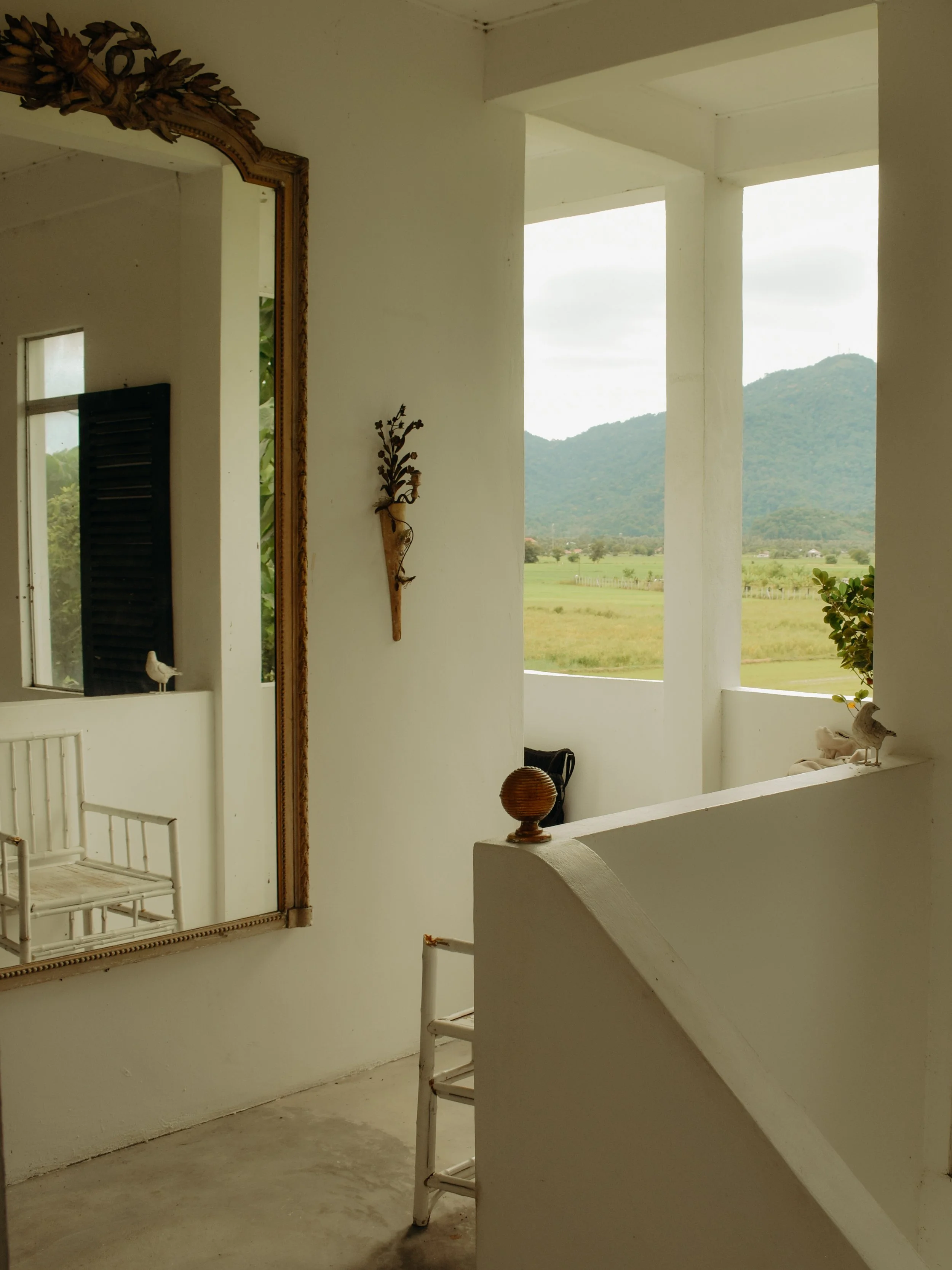 A section of an interior with white walls and a large window or opening showing a mountainous landscape in the distance. There are a decorative mirror, wall-mounted ornament, and some furniture.
