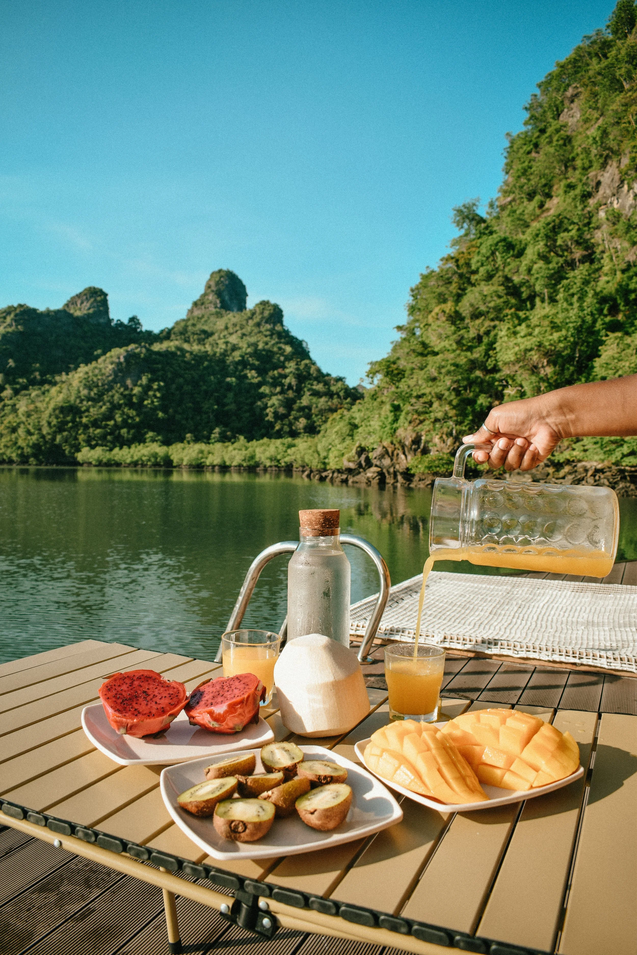 A lakeside breakfast spread featuring fresh fruits such as cut cantaloupe, dragon fruit, sliced kiwis, and a plate of sliced papaya, with glasses of orange juice on a wooden table, and a hand pouring juice from a glass pitcher. In the background, there's a body of water with lush green forested hills and a clear blue sky.