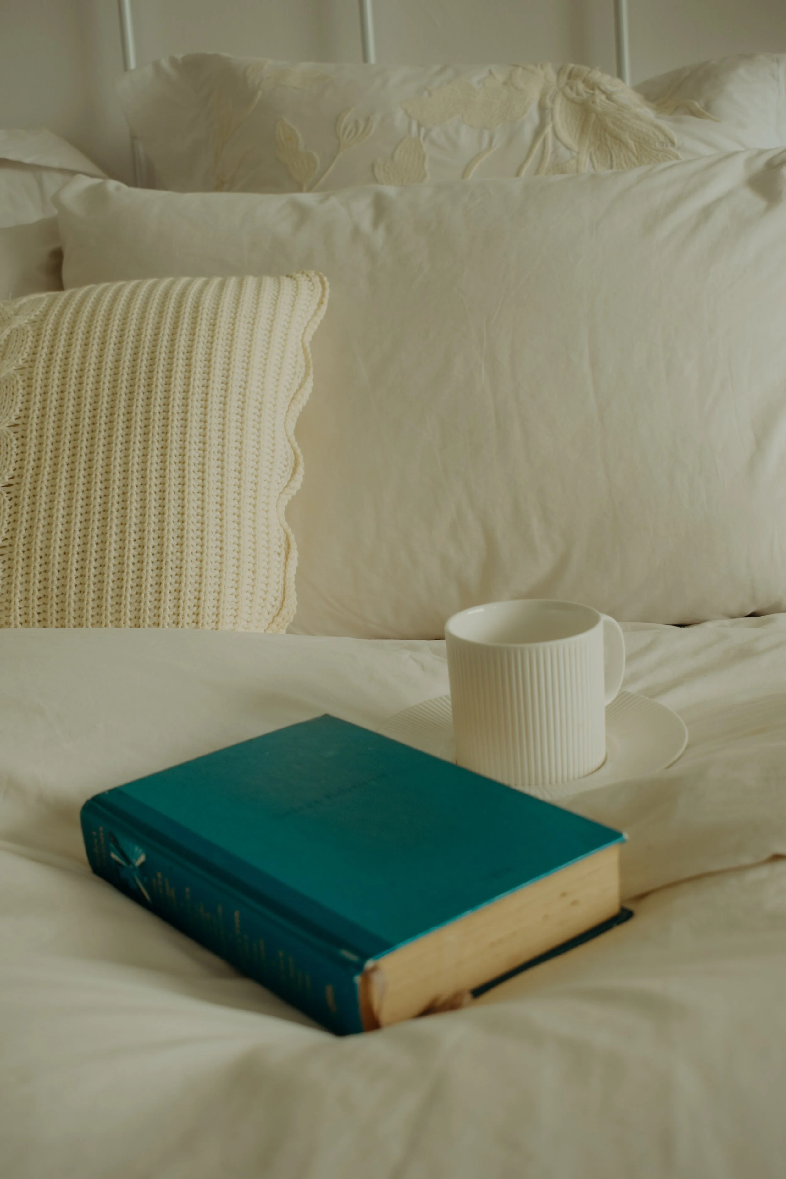 A bed with cream-colored pillows, a blue book, a white mug, and a white knitted pillow with lace detailing.
