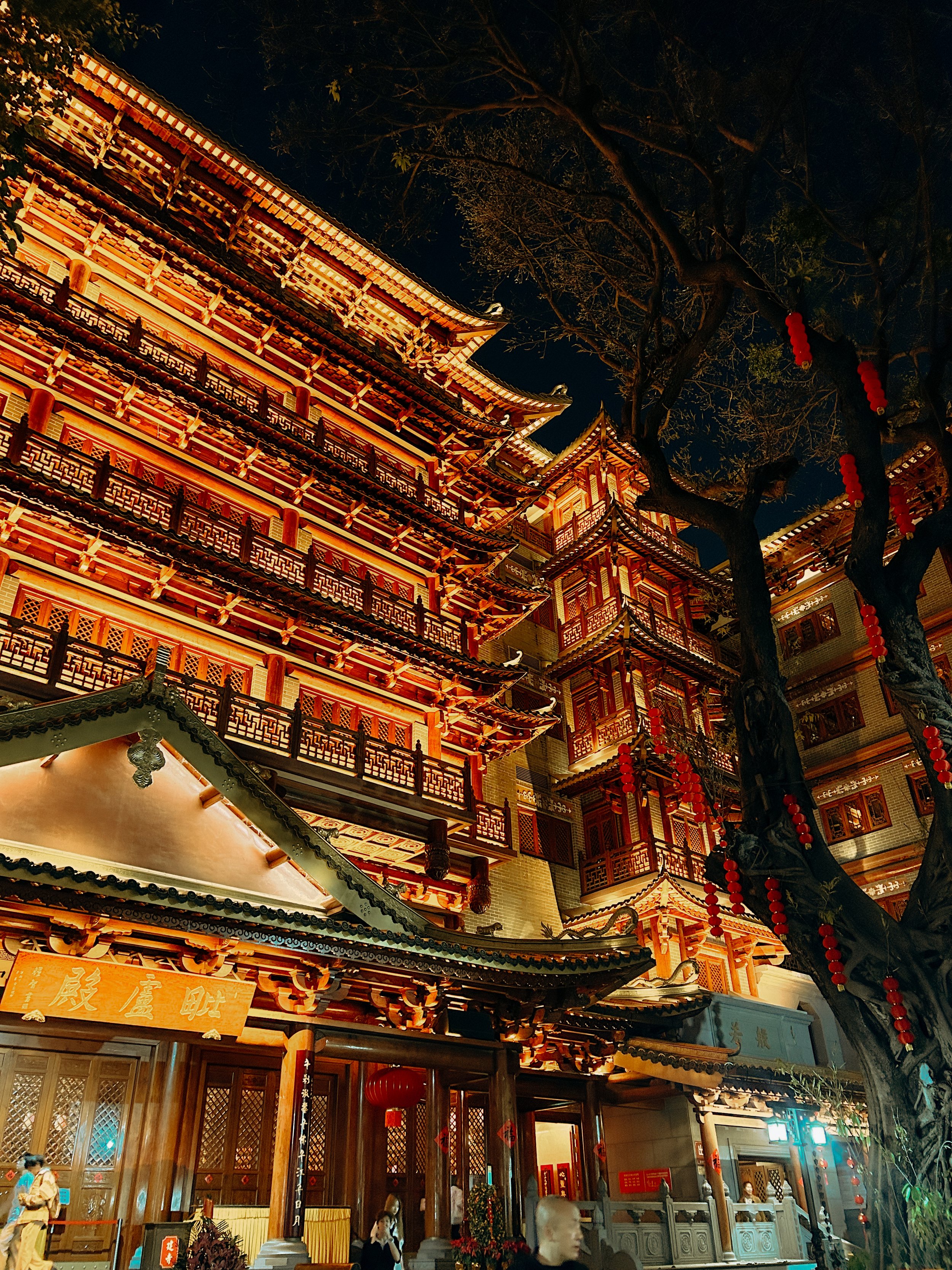 Night view of a traditional Chinese temple with ornate wooden architecture, detailed roof ridges, and hanging red lanterns. The temple is illuminated with warm lighting, and there are people walking and standing in front of it.