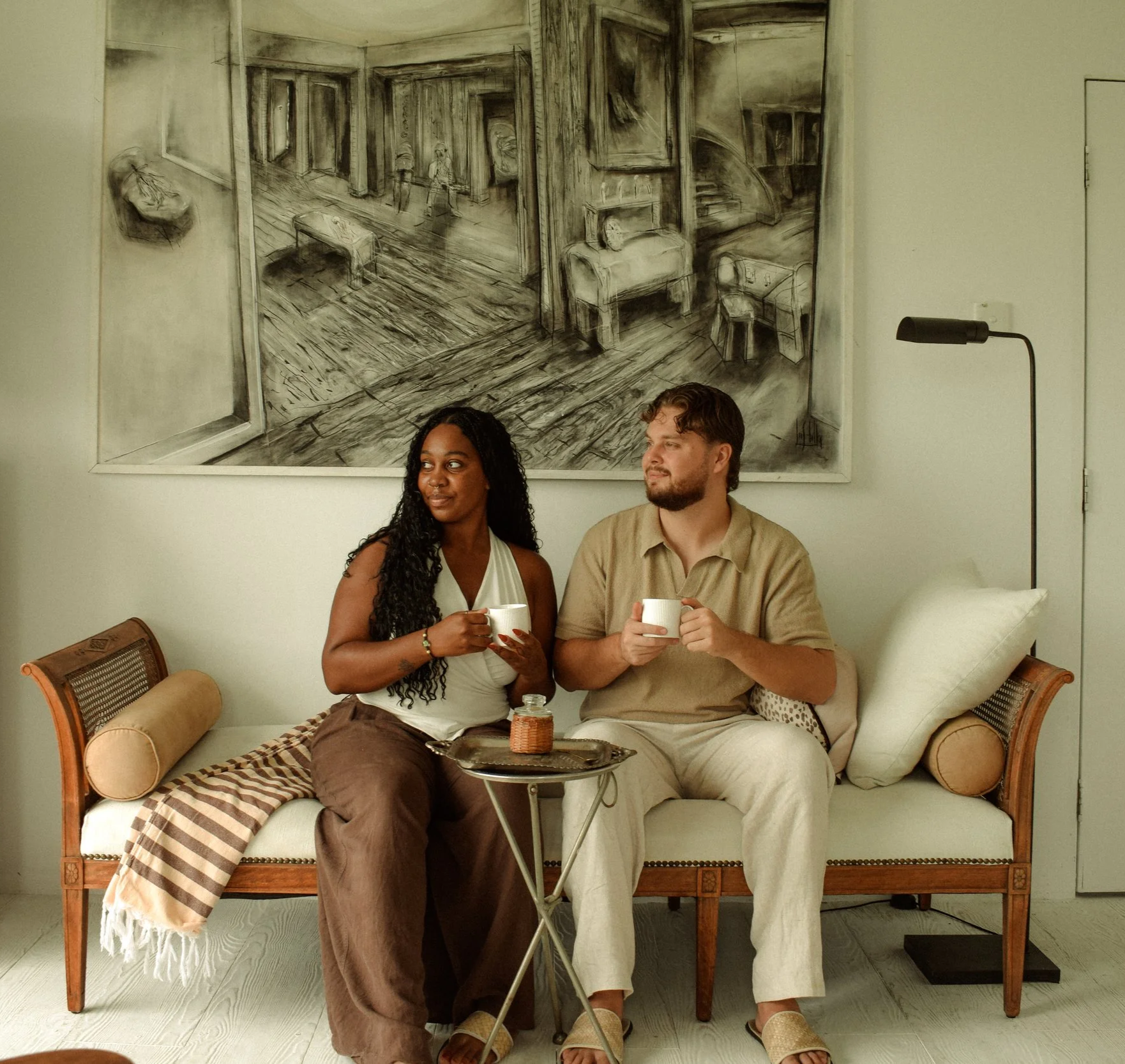 Two people sitting on a cream-colored sofa with patterned cushions, holding white mugs, in front of a black and white painting of an interior scene, with a small side table with a wire basket and a small silver tray. A woman has long curly hair and is wearing a sleeveless white top and brown pants. A man has short curly hair and a beard, and is wearing a beige shirt and cream-colored pants. A black lamp with a curved arm is on the right side of the image.