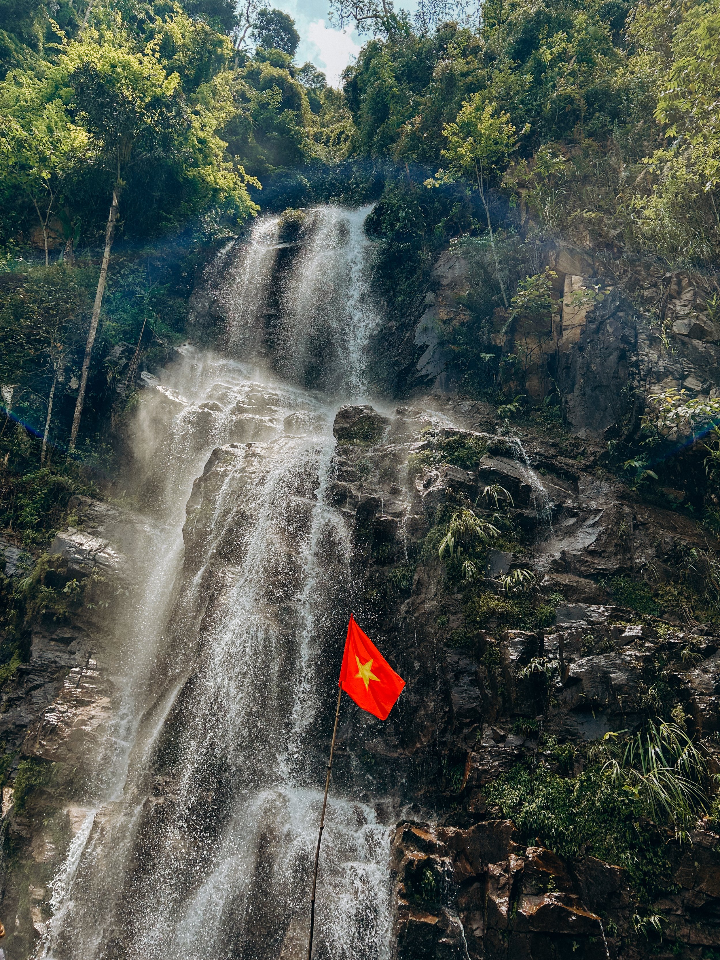 A waterfall cascading down dark rocks in a lush green forest, with a small Vietnamese flag on a pole at the bottom of the waterfall.