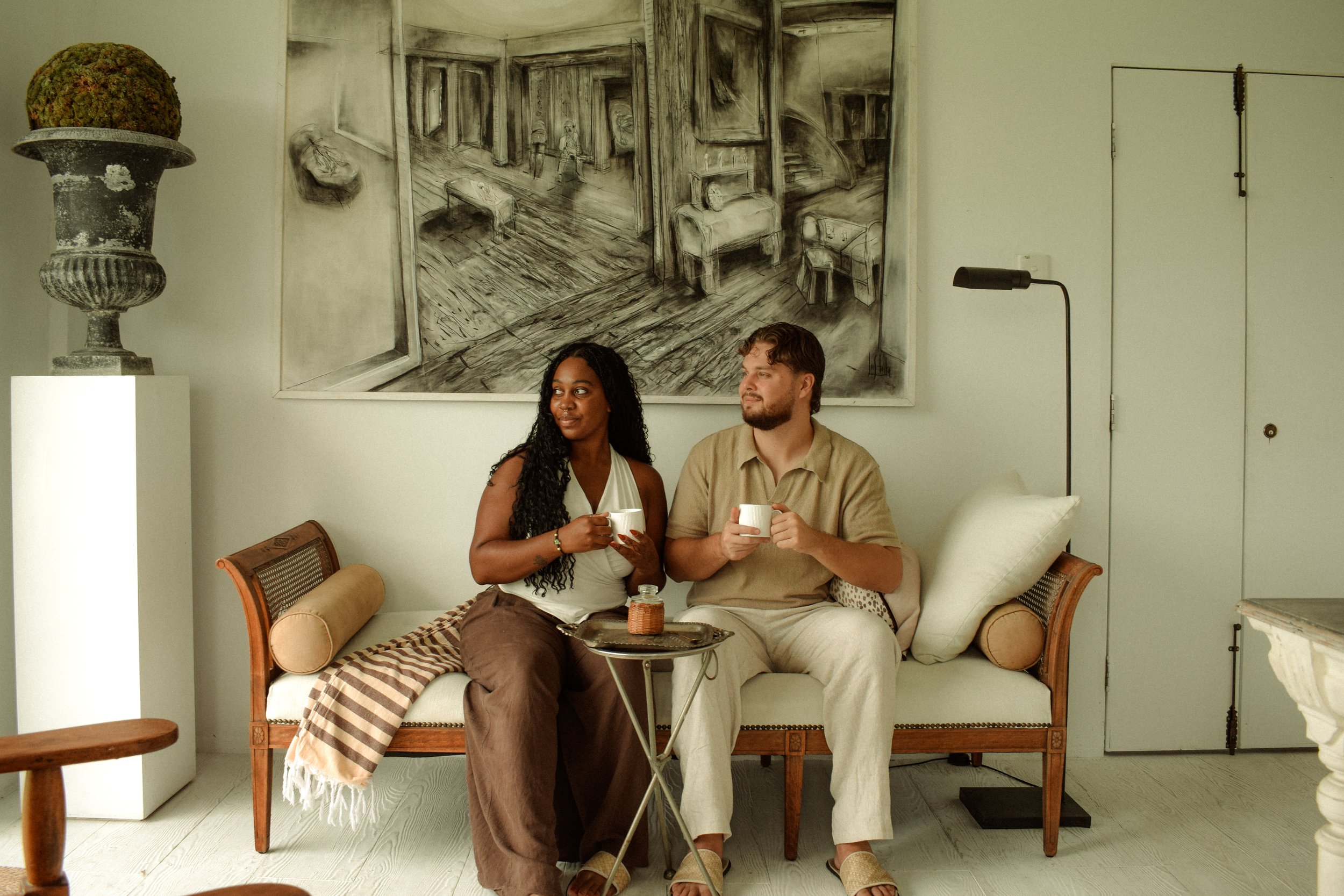 Two people sitting on a white sofa, holding cups, with a large black and white artwork behind them depicting an interior scene. A woman with dark curly hair is wearing a white top and brown pants, while a man with light brown hair and a beard is wearing a beige shirt and light-colored pants.
