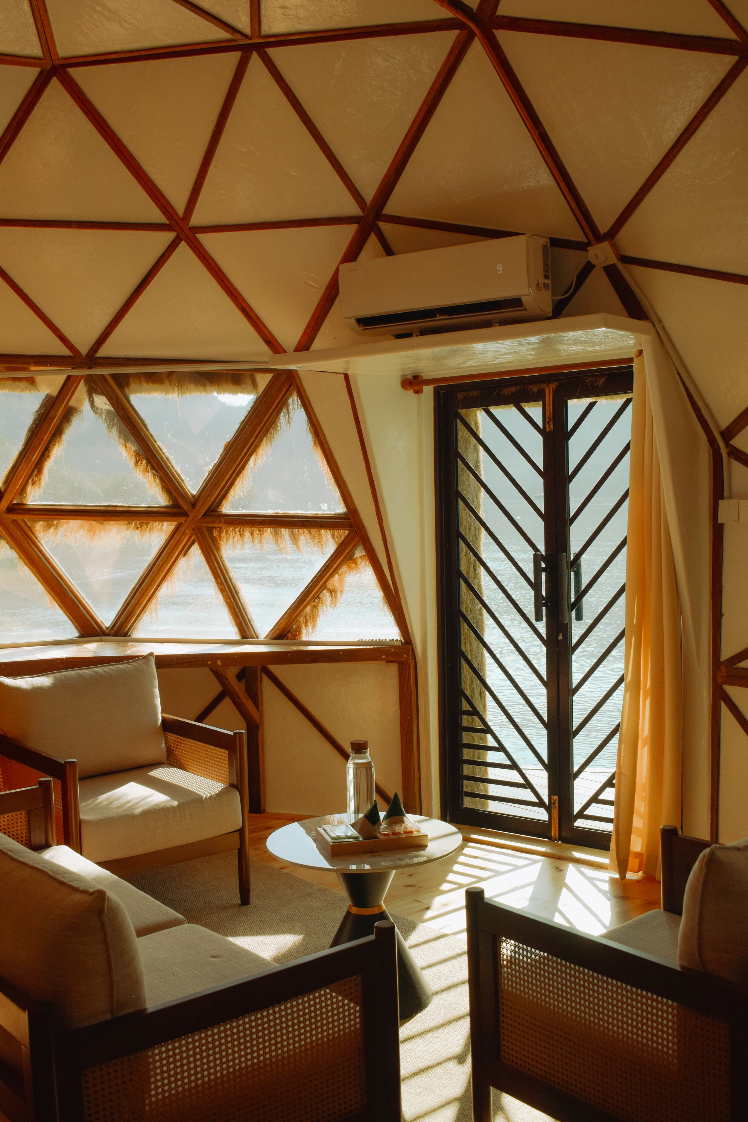 Interior of a room with wooden geometric patterned walls and a view of water outside through a glass door with metal bars. There is a cream-colored couch, a small round coffee table with books and a water bottle, and yellow curtains.