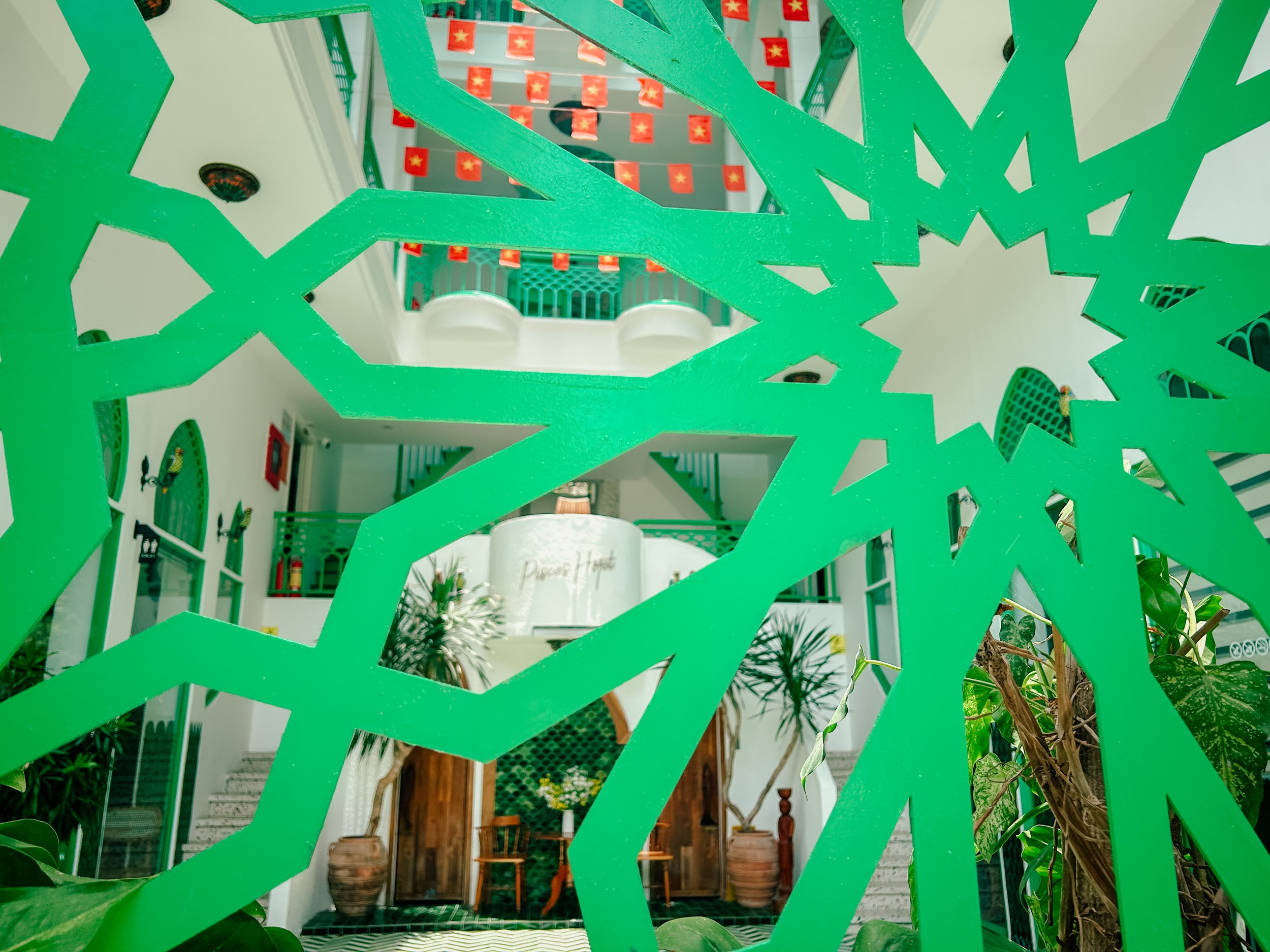 View of an interior space with a staircase and seating area, seen through a decorative green wooden lattice with geometric cutouts and several small red flags with yellow stars hanging from the ceiling.