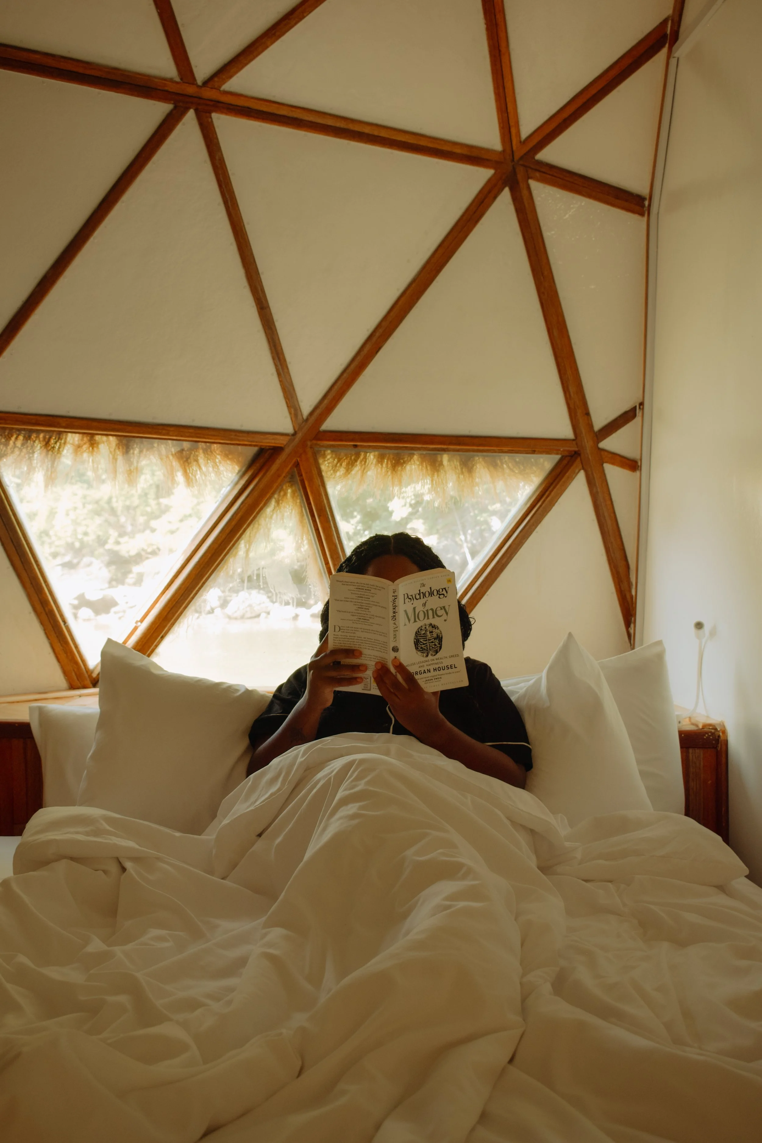 A person sitting in bed reading a book titled 'The Psychology of Money' in a room with a wooden geodesic dome ceiling and a window showing trees outside.