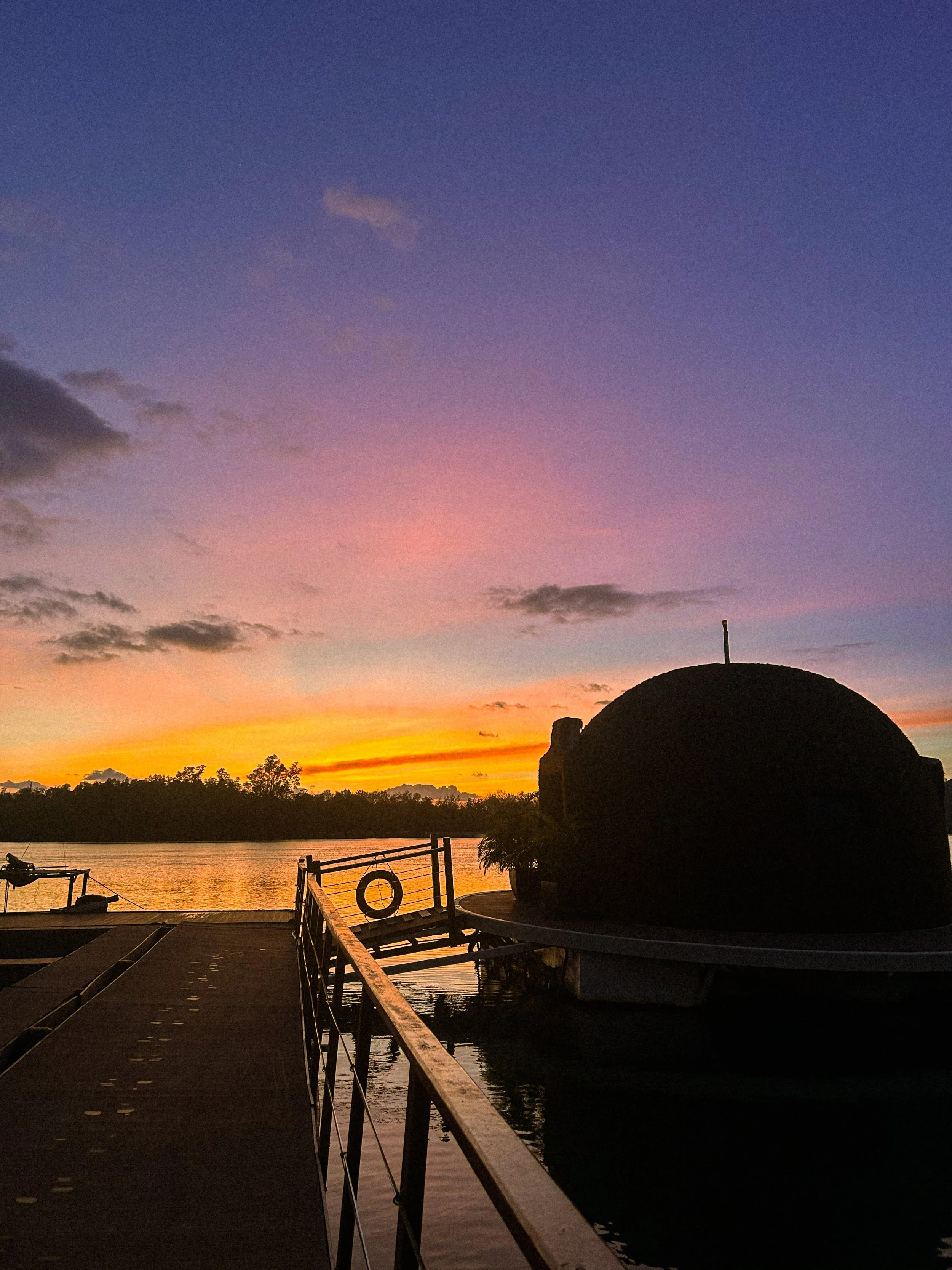 A sunset scene by the water with a silhouette of a domed structure on the right and a railing along a dock leading into the water, with trees and a colorful sky in the background.