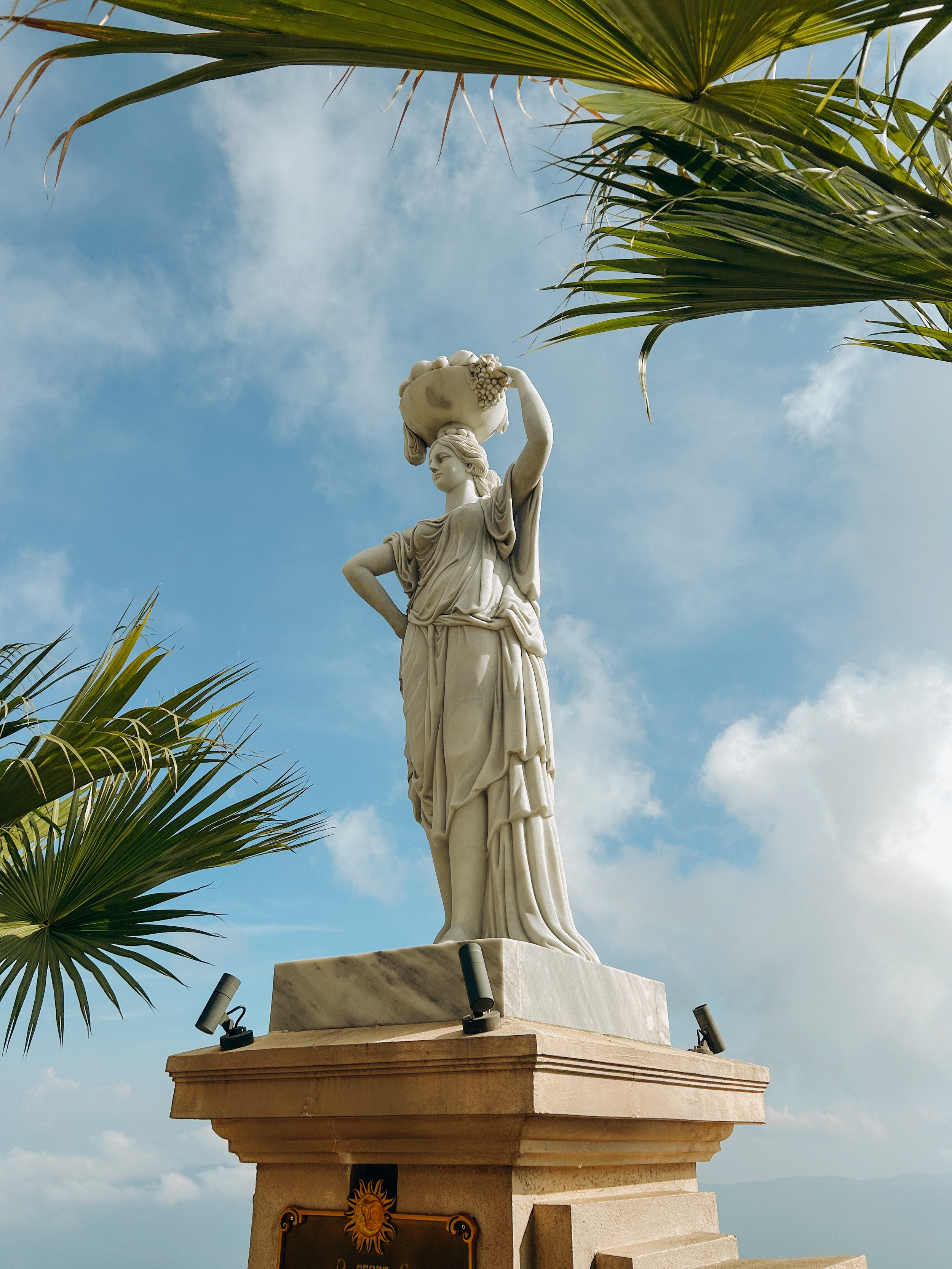 Statue of a woman holding a basket of grapes, with palm fronds in the foreground and a cloudy sky in the background.