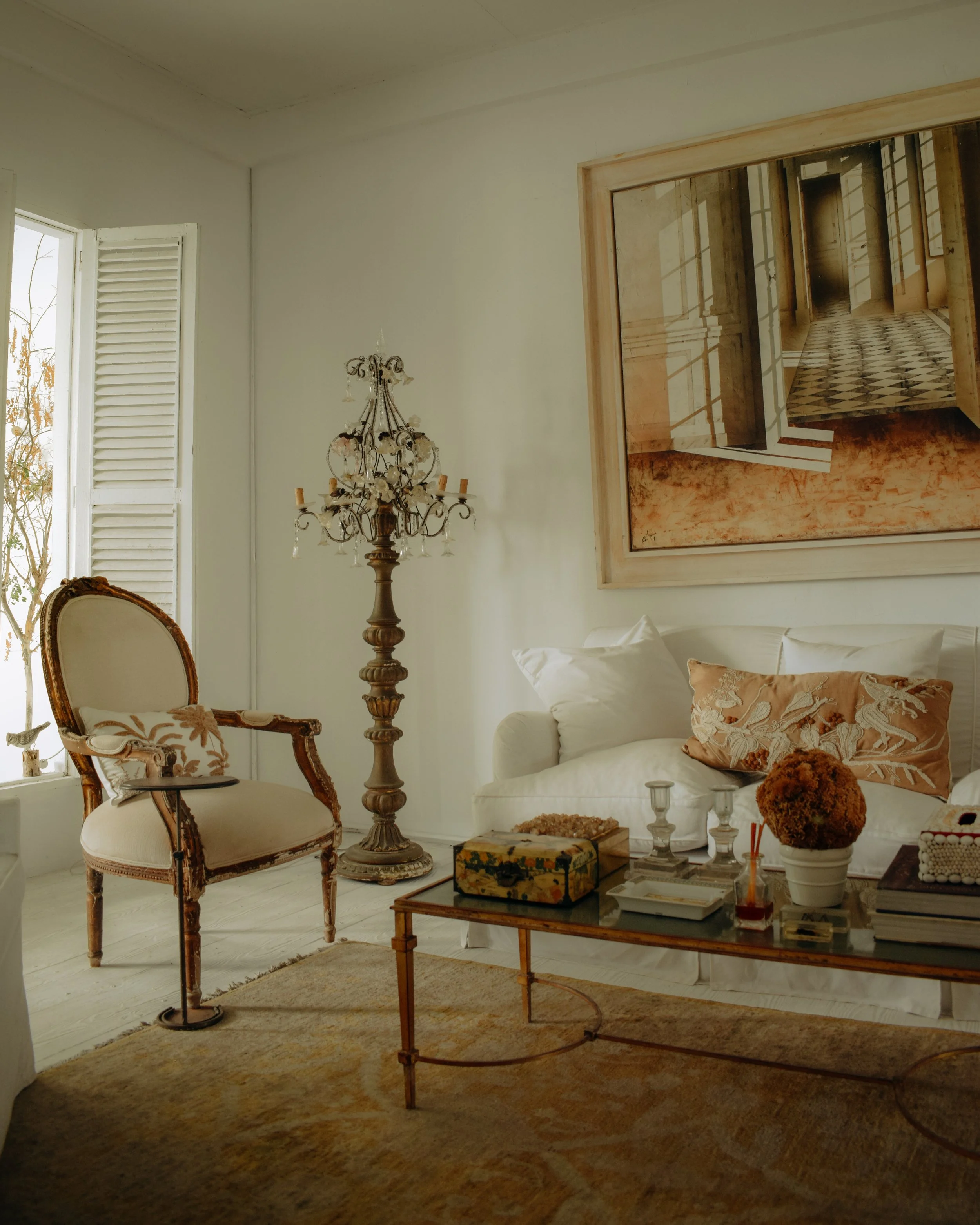 A living room with a large framed photo on the wall, a tall ornate chandelier, a vintage armchair, a white sofa with decorative pillows, a coffee table with various items including a box and glass cups, and a window with white shutters. The room has wooden flooring and a beige rug.