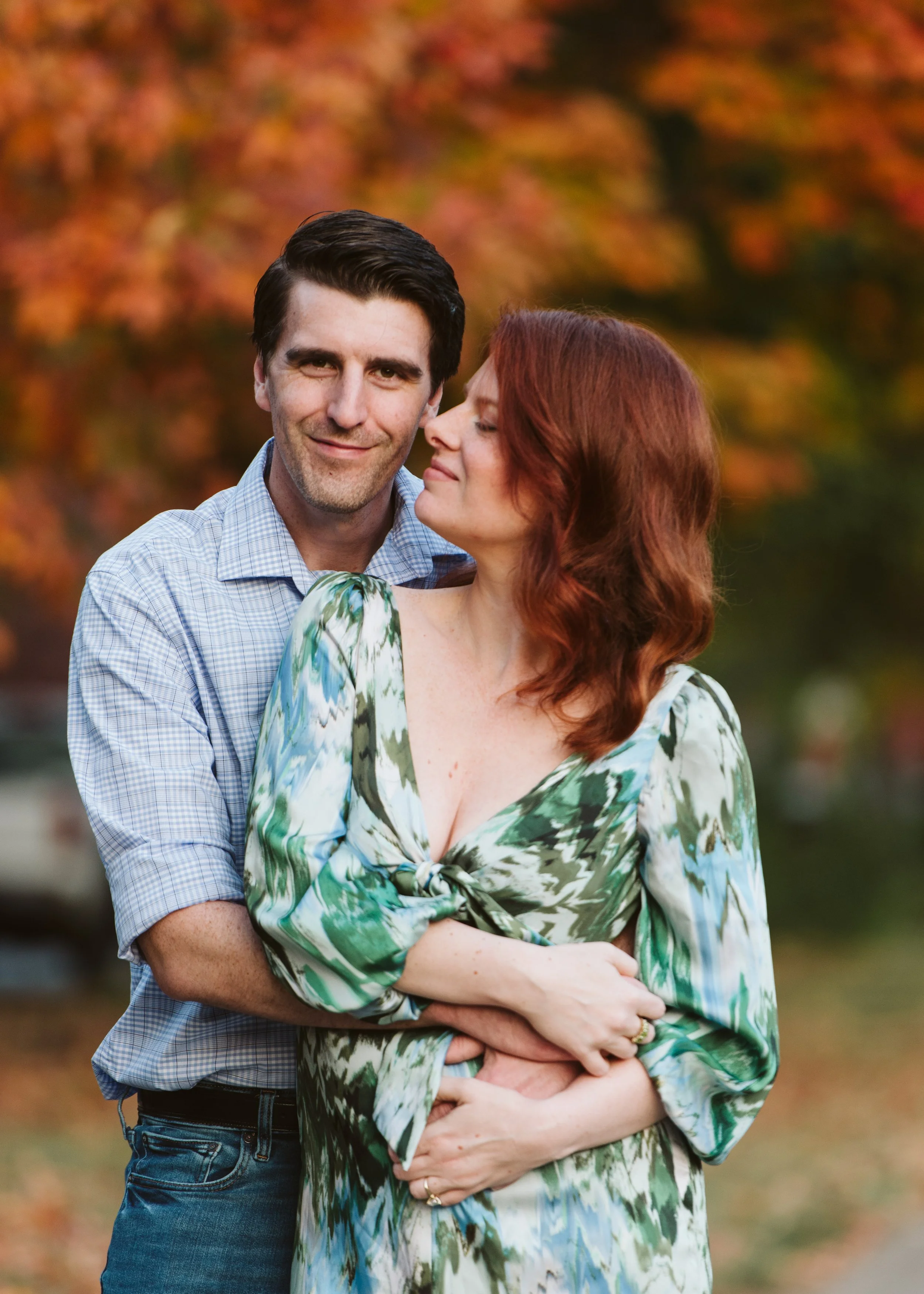 A couple stands outdoors with autumn foliage in the background. The man has dark hair and is smiling, while the woman with red hair is facing closer to him with her eyes closed and a content expression. They are embracing each other, with the woman's arms crossed in front of her.