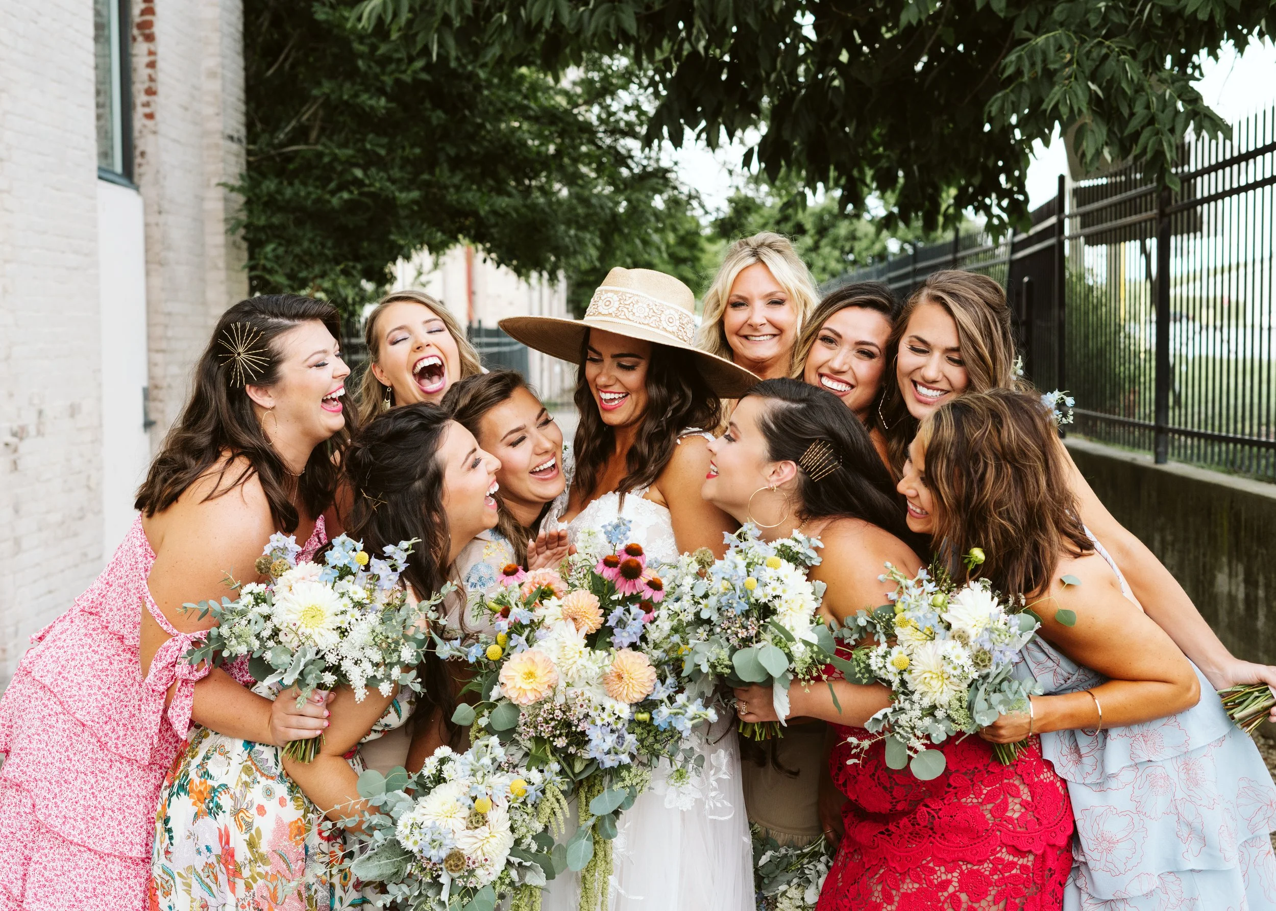 A group of women hugging and smiling, celebrating outdoors, with a bride holding a large bouquet of white and pastel flowers, and a woman in a wide-brimmed hat. They are dressed in colorful dresses, in a setting with trees and a fence.