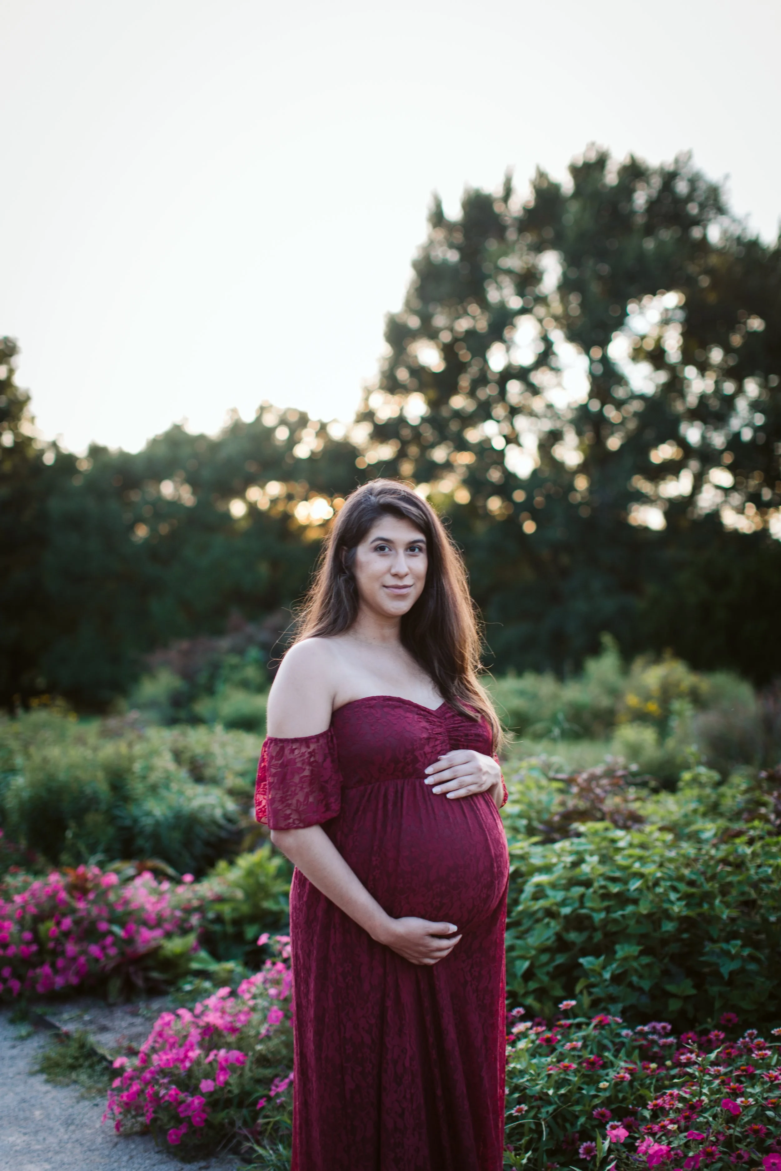 A pregnant woman in a strapless burgundy lace dress stands in a garden with pink flowers and green foliage. She cradles her belly with one hand and looks into the camera with a gentle smile, with trees and the setting sun in the background.
