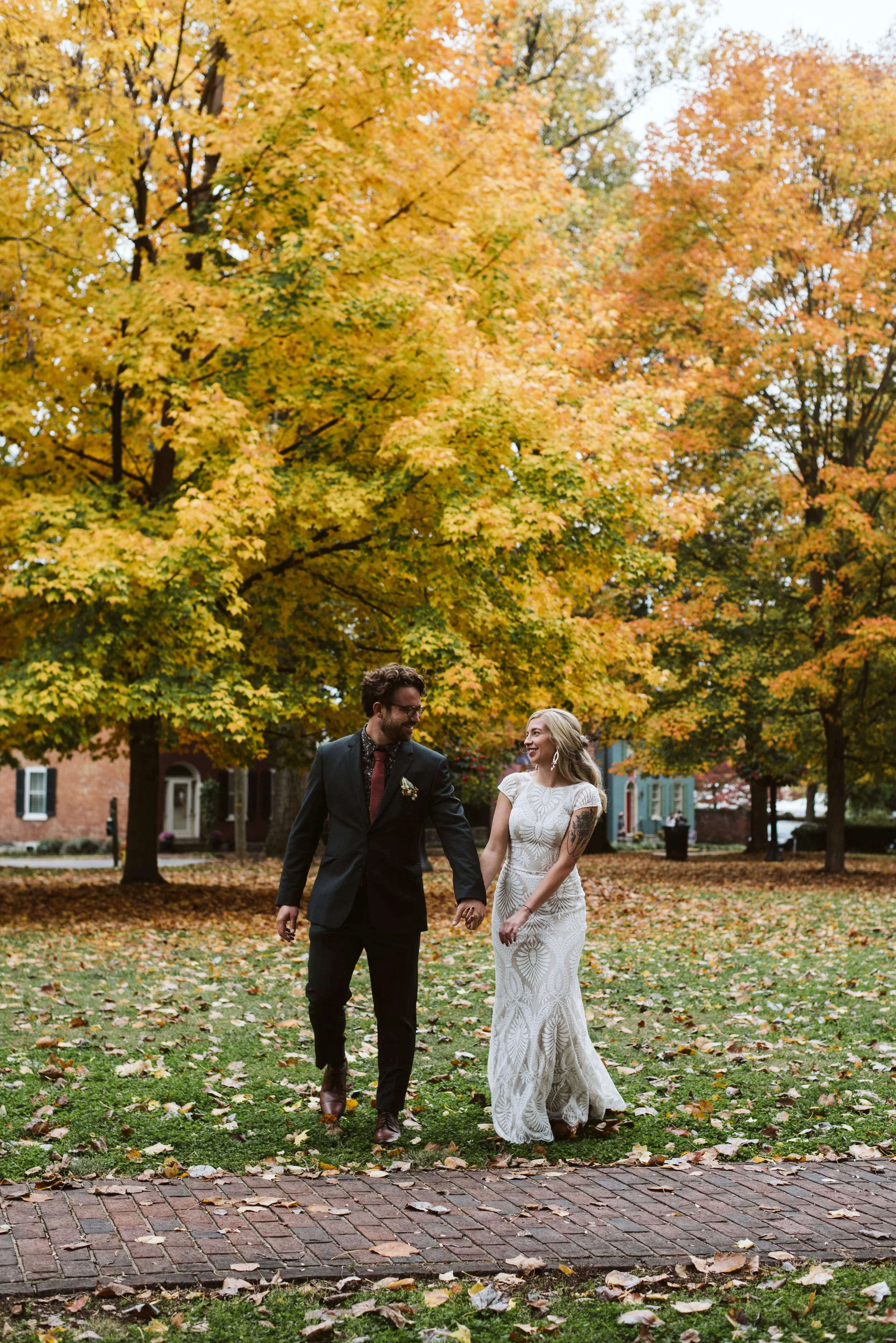 A couple, a man in a black suit and a woman in a white lace dress, walking hand in hand in a park with fall foliage, with trees and brick buildings in the background, during autumn.