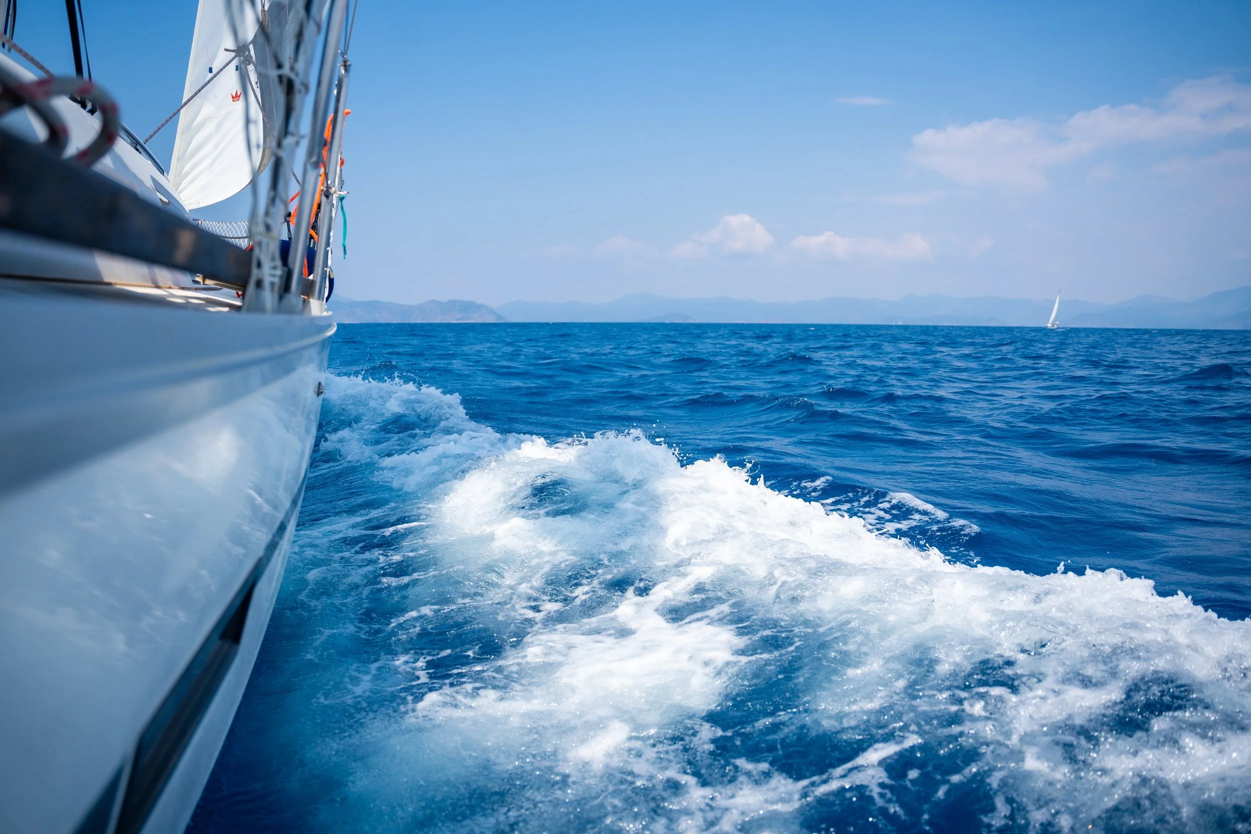 Un'imbarcazione a vela che si muove sull'oceano con un'altra vela visibile in lontananza e le montagne sullo sfondo.